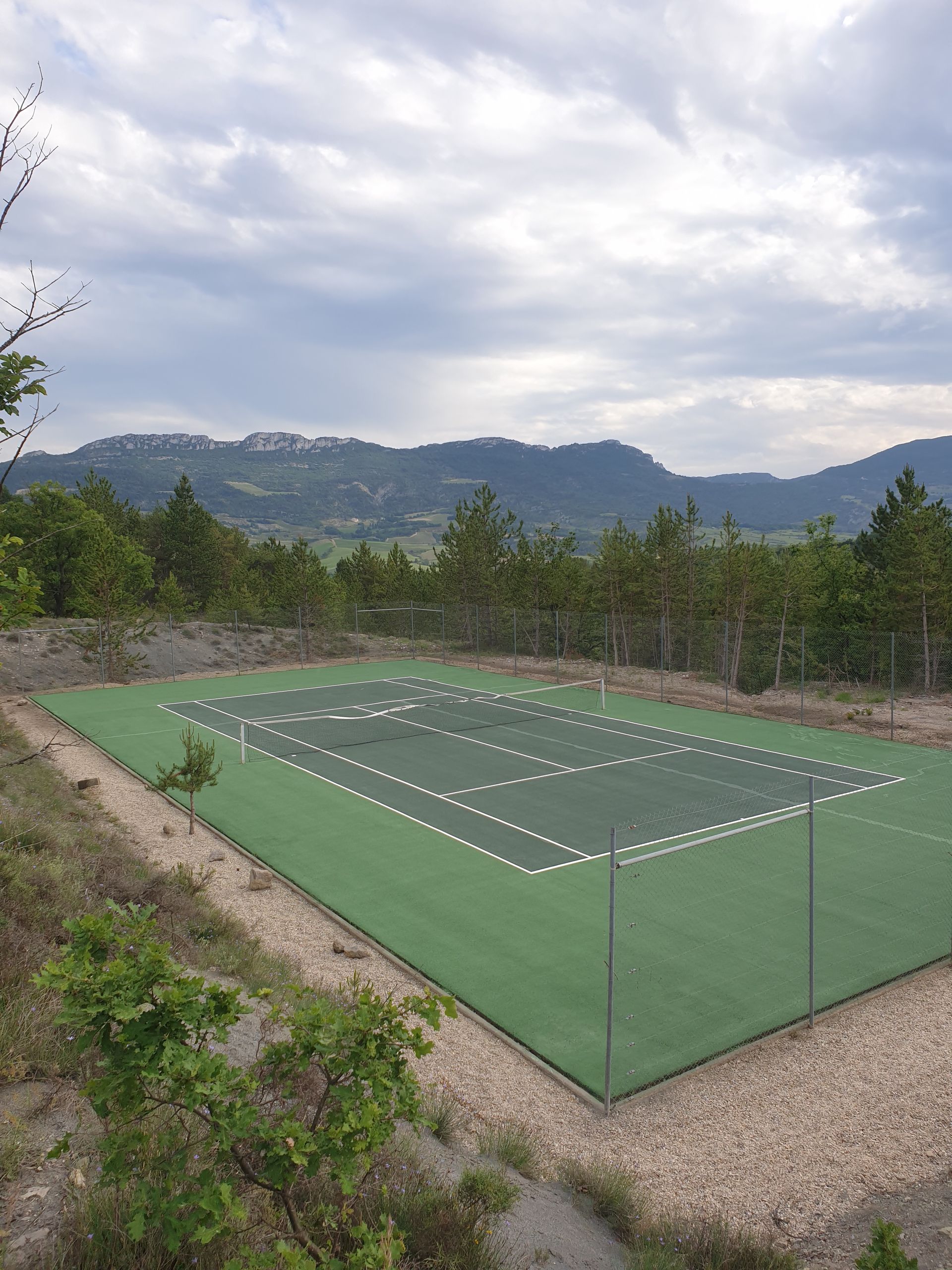 Court de tennis extérieur vert, clôturé, avec des arbres et des collines au loin sous un ciel nuageux.