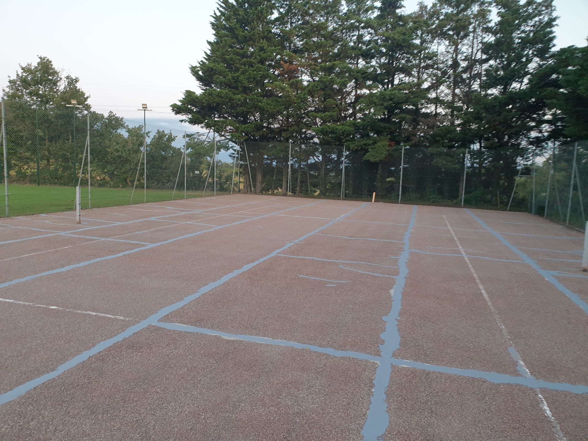Un terrain de sport vide en gravier, avec des lignes blanches, entouré d'une clôture et d'arbres.