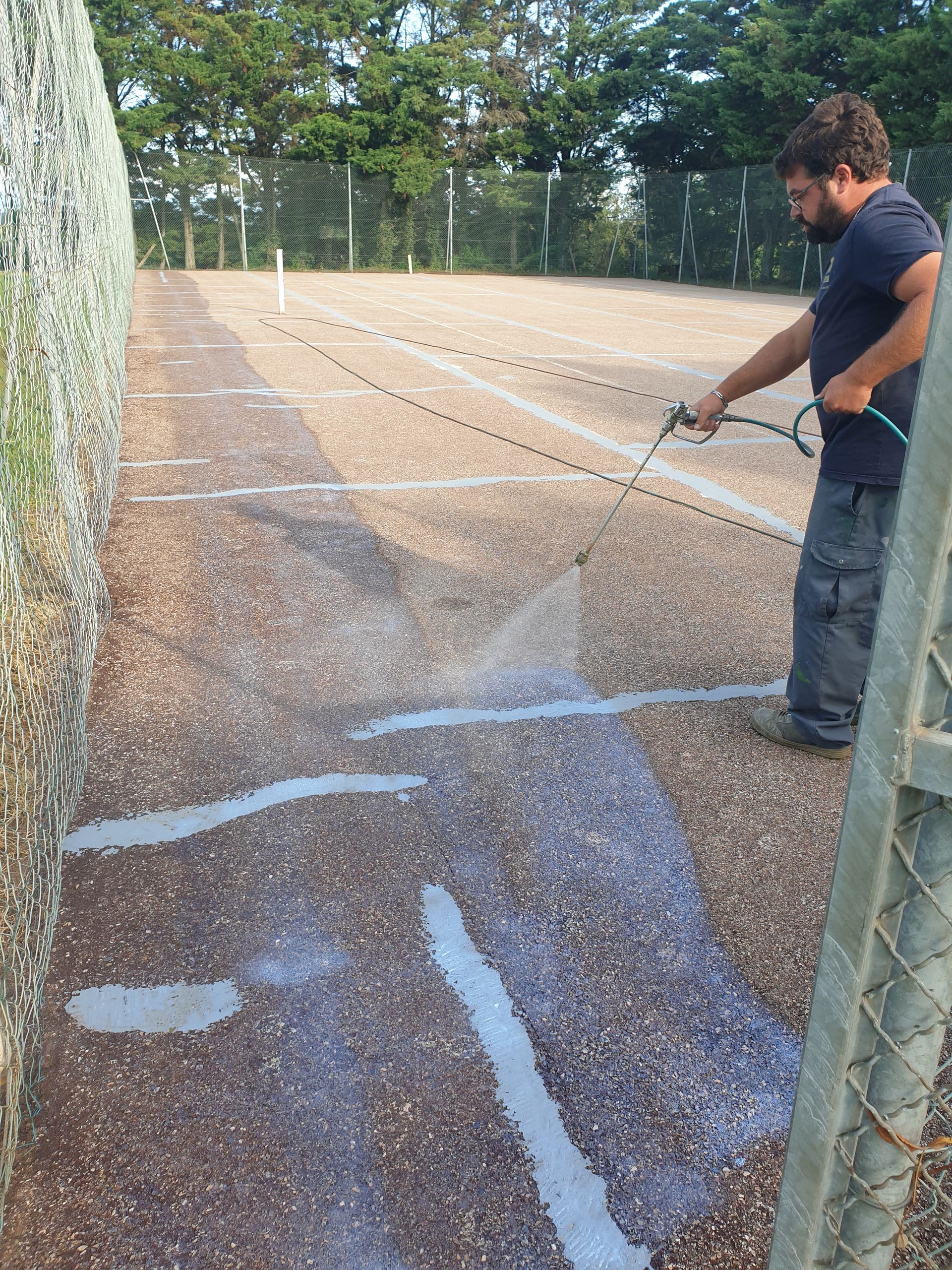 Un homme arrose un court de tennis, près d'une clôture, par une journée ensoleillée.