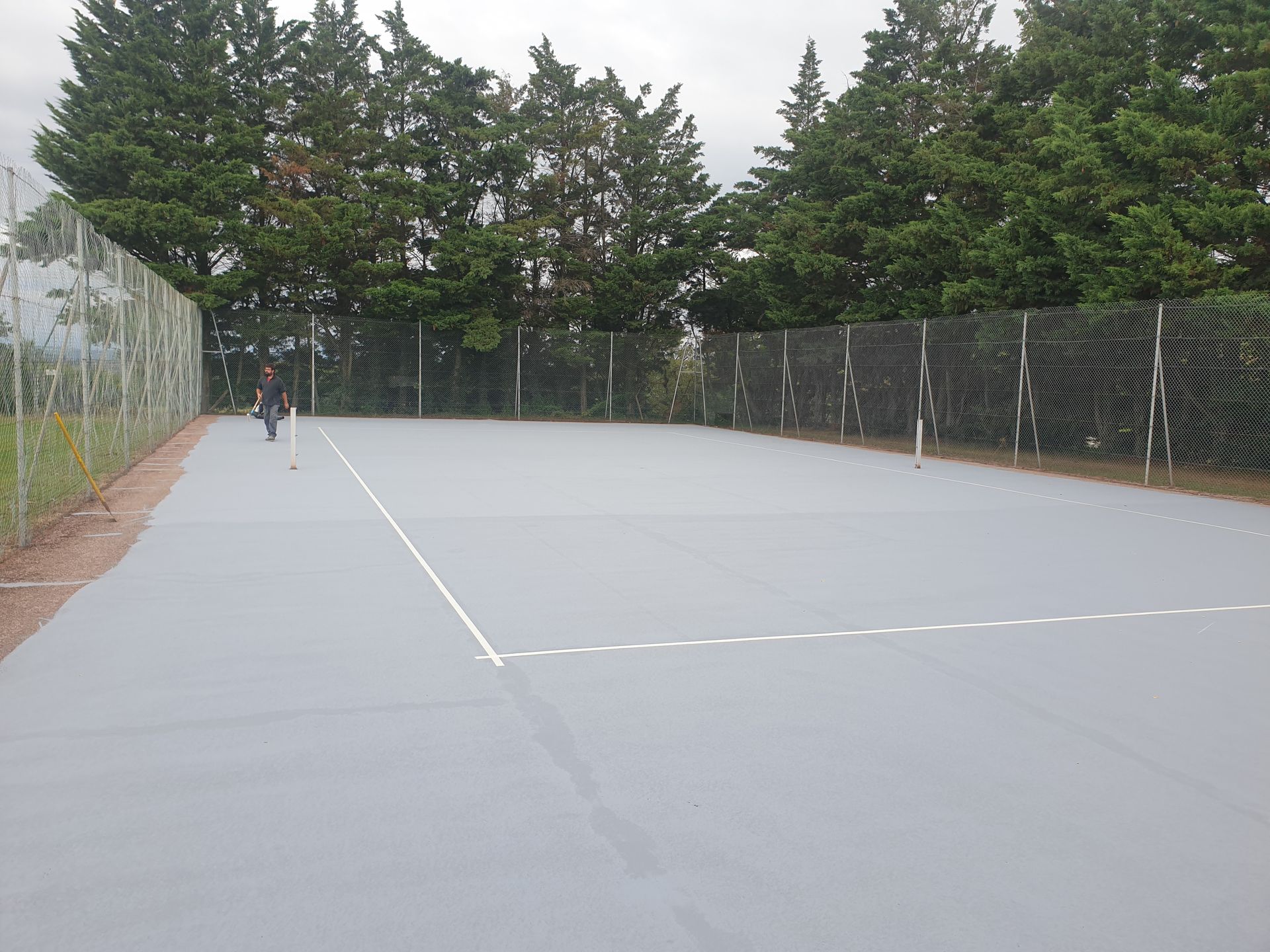 Terrain de handball gris avec des lignes blanches, clôturé sur deux côtés, arbres en arrière-plan.