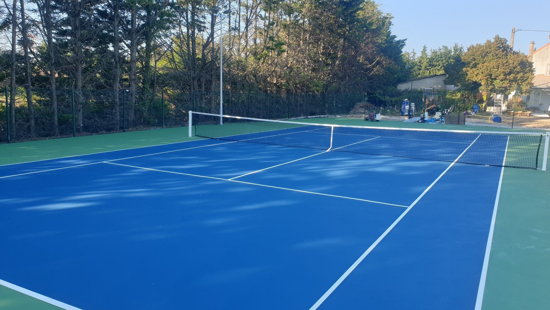 Court de tennis bleu avec lignes et filet blancs, entouré d'une surface verte et d'arbres.