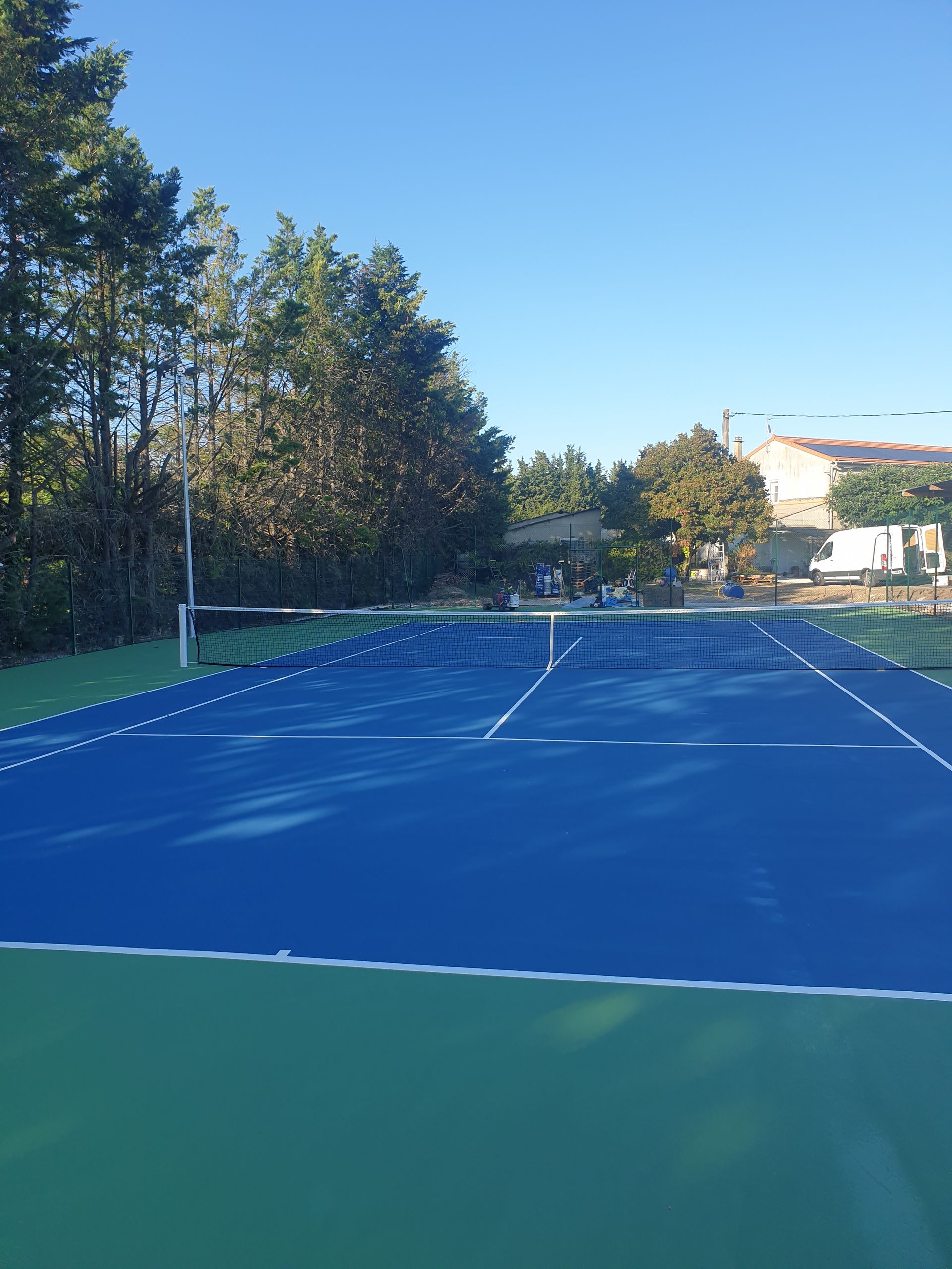 Court de tennis bleu et vert avec lignes blanches, filet et arbres sur fond de ciel bleu.