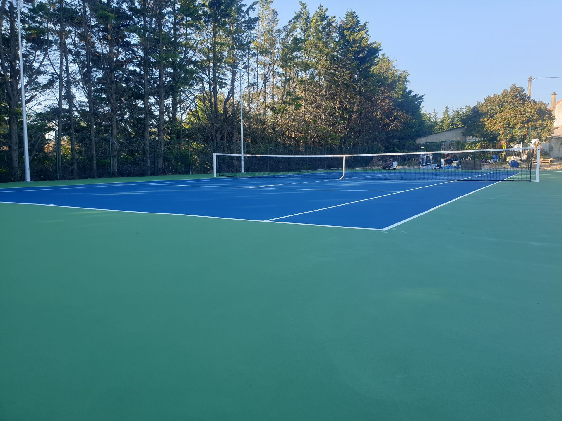 Court de tennis avec surface de jeu bleue, espace vert environnant et filet. Arbres en arrière-plan.
