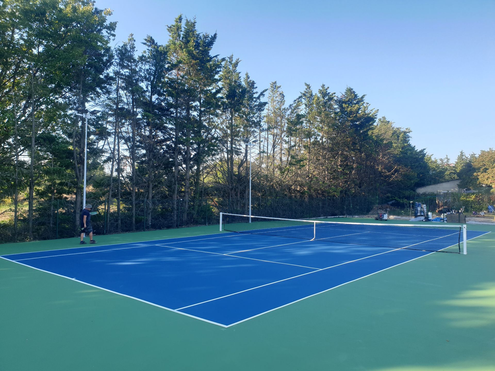 Court de tennis bleu avec lignes blanches, espace vert environnant, arbres et une personne jouant.