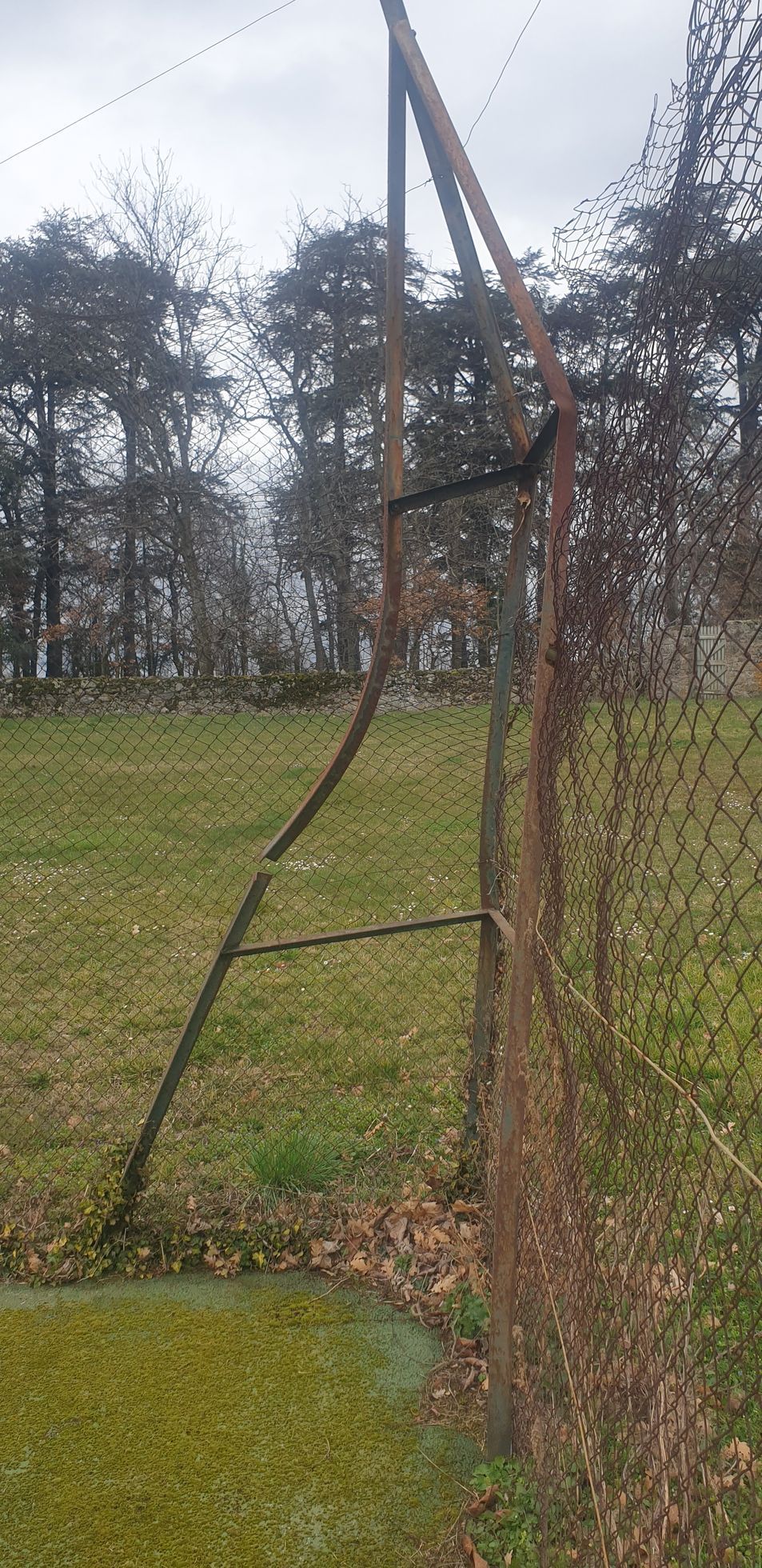 Un poteau de clôture rouillé et un grillage dans un champ herbeux avec des arbres en arrière-plan, sous un ciel nuageux.