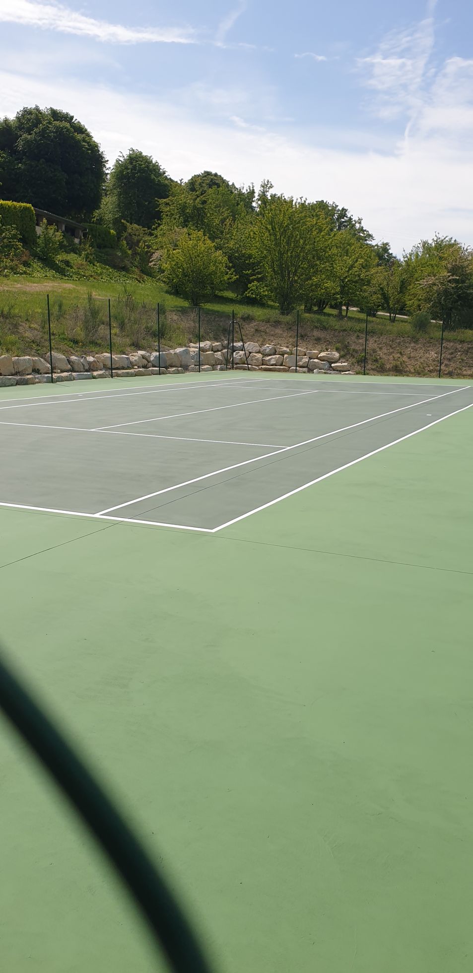 Court de tennis avec surface verte et grise, lignes blanches, arbres en arrière-plan, ciel bleu.