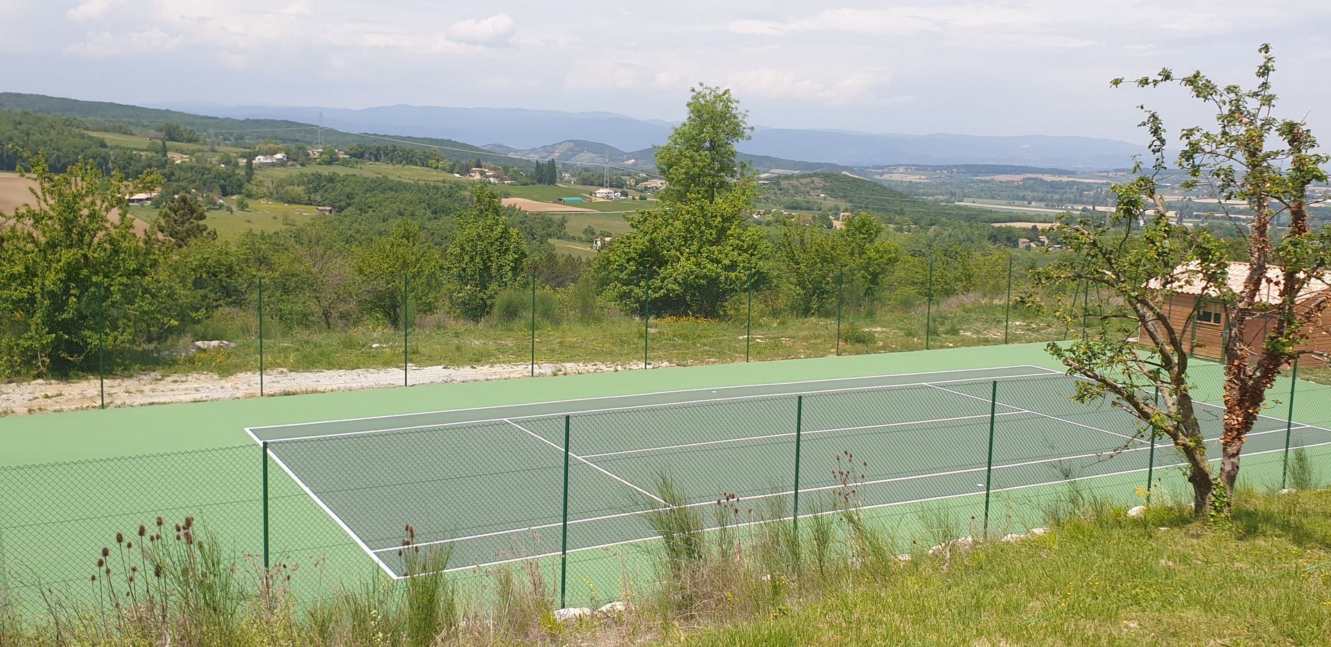 Court de tennis vert avec clôture en fil de fer, arbres et collines ondulantes en arrière-plan par une journée ensoleillée.