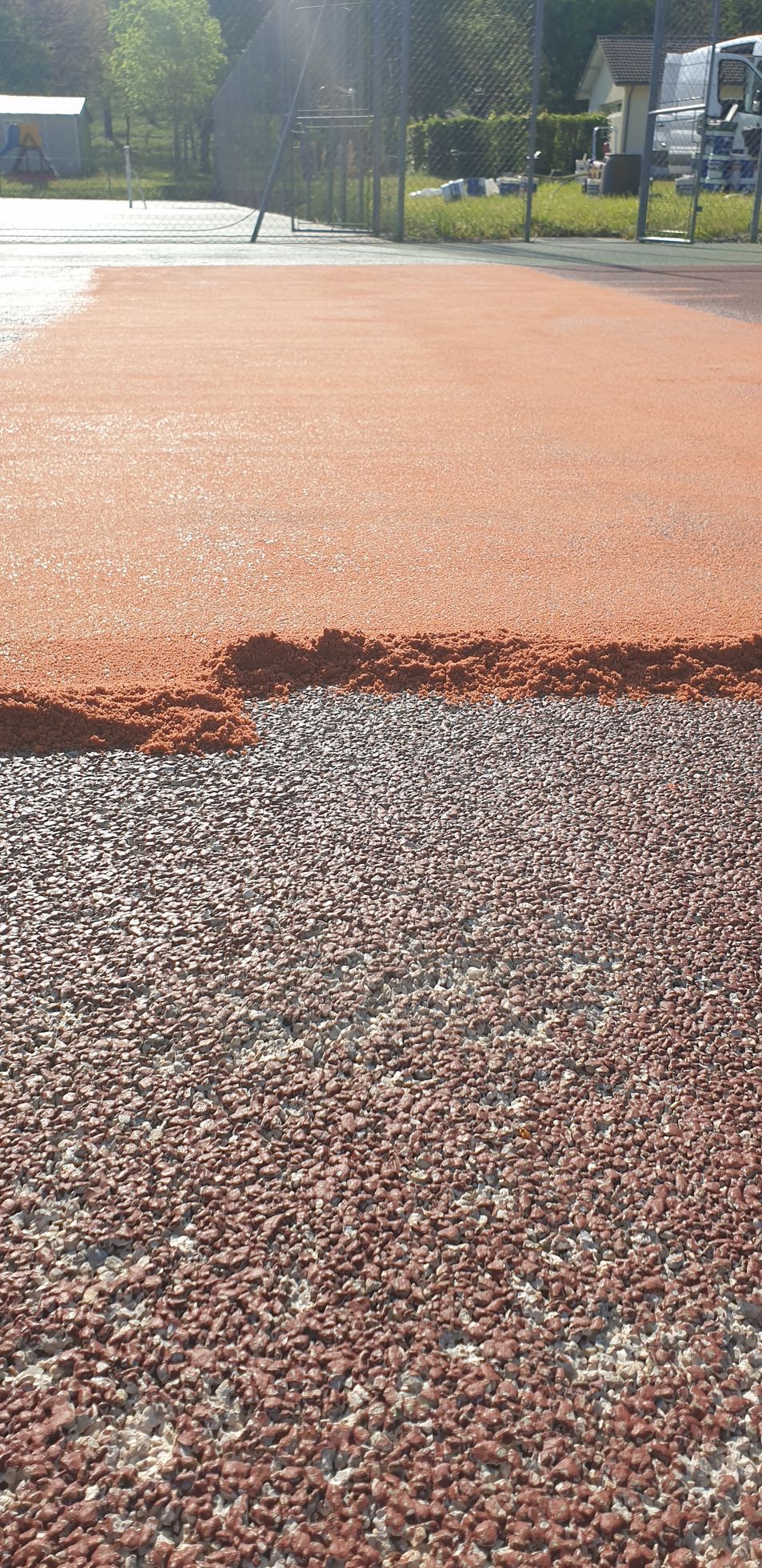Une surface brun rougeâtre laisse place à une texture de gravier plus foncée, probablement un terrain de sport en construction.