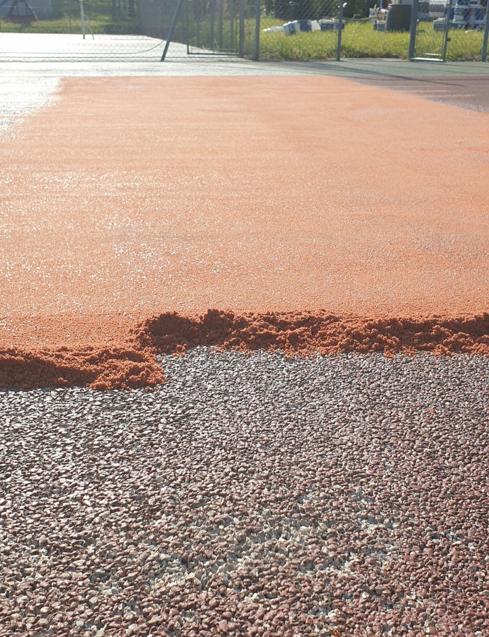 Travaux de réfection d'un court de tennis en cours ; application d'une couche supérieure rouge-orange sur une couche de gravier fin.