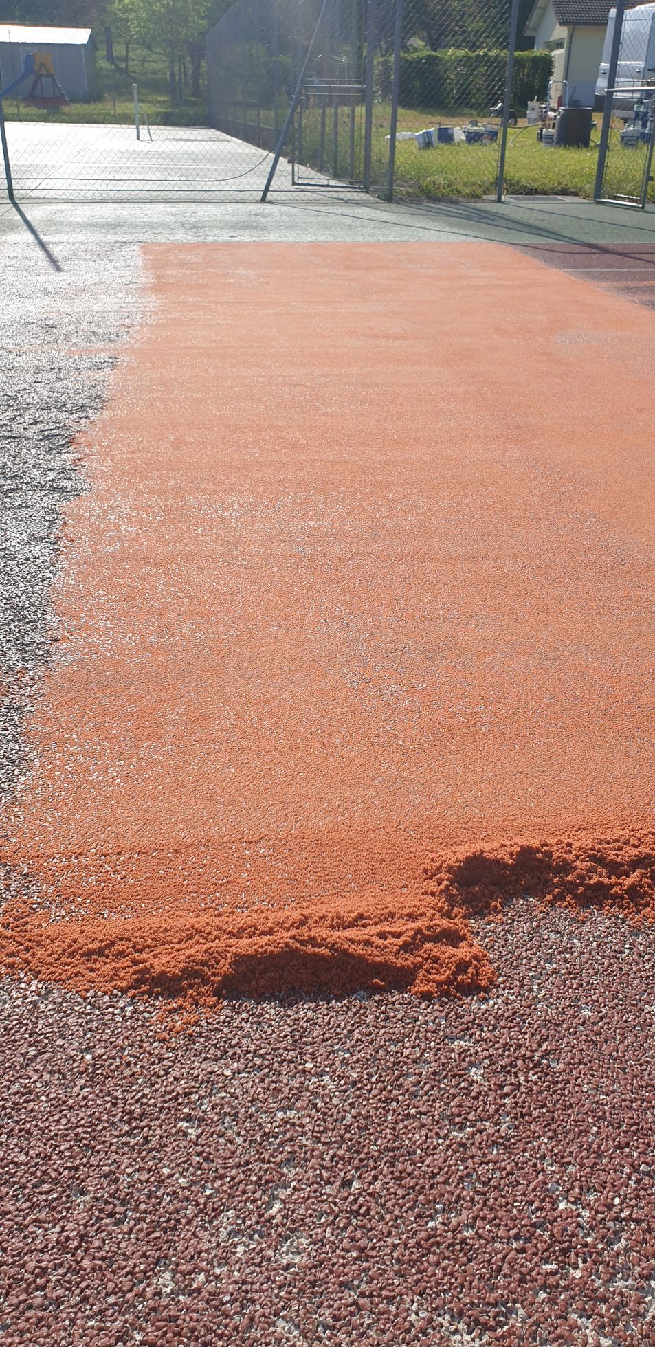 Un court de tennis en terre battue avec une section en terre battue rouge fraîchement posée, entouré d'une clôture et de verdure.