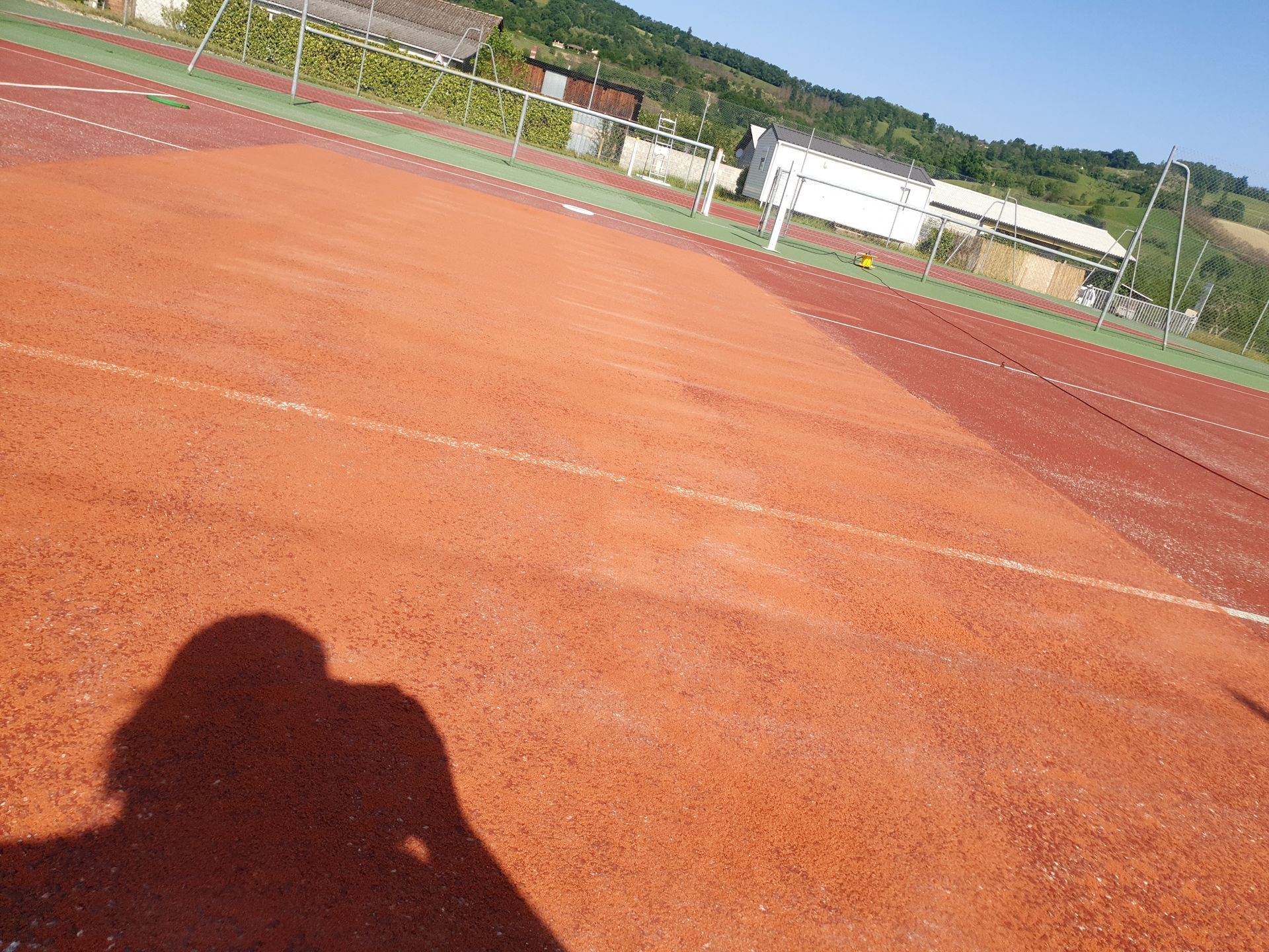 Un terrain de sport rouge avec des lignes blanches et l'ombre d'une personne. À l'arrière-plan, des arbres et des bâtiments.
