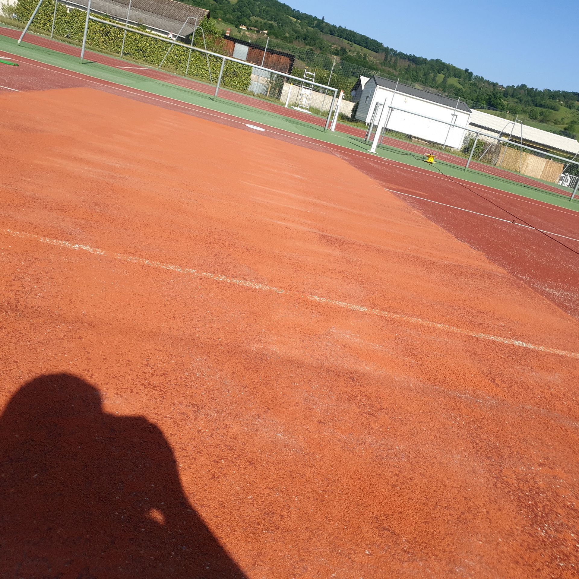 Terrain de sport en terre battue rouge avec des lignes blanches. Ombre d'une personne au premier plan. Bâtiments et arbres à l'arrière-plan.