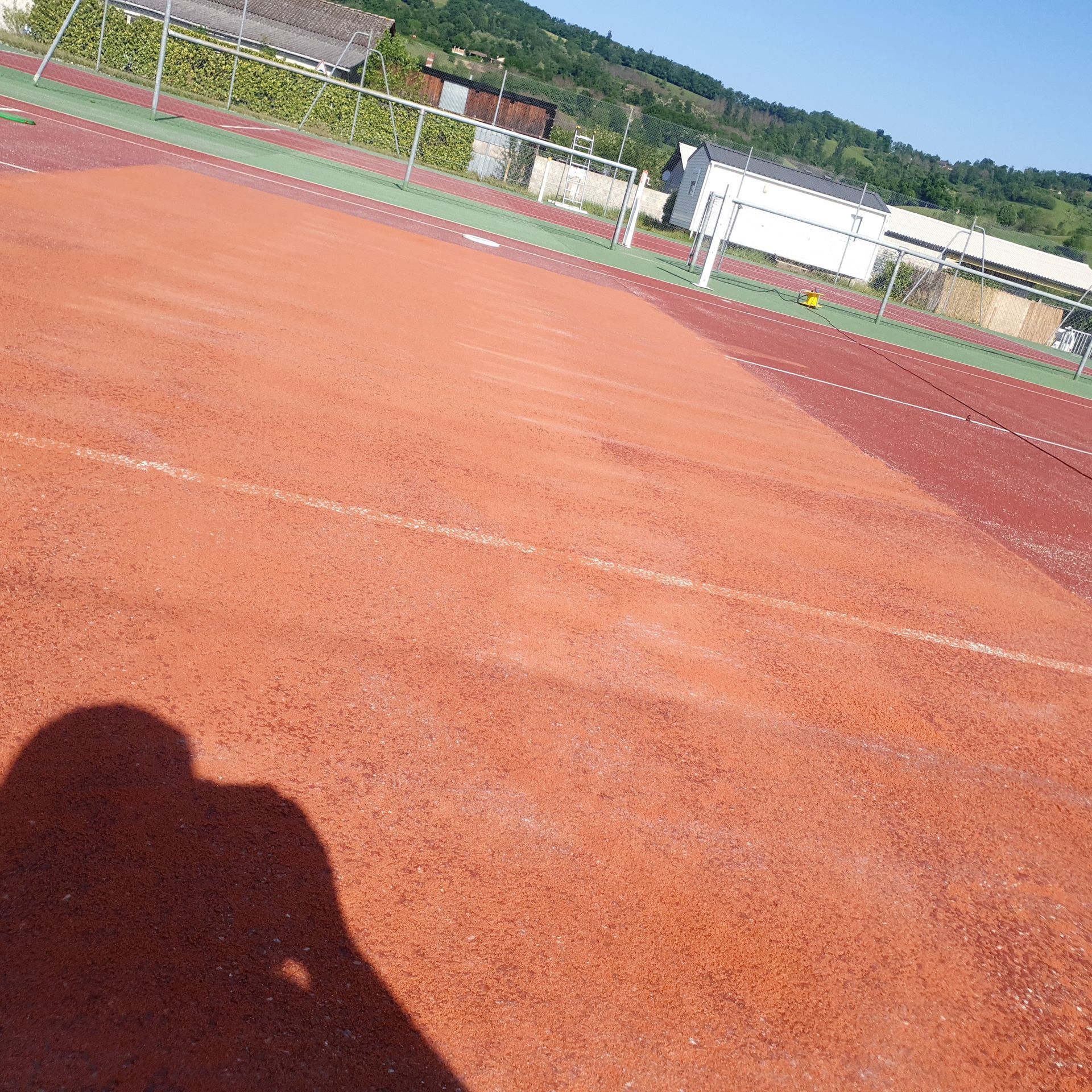 Terrain de sport rouge avec lignes blanches, ombre d'une personne. Bâtiments et arbres en arrière-plan.
