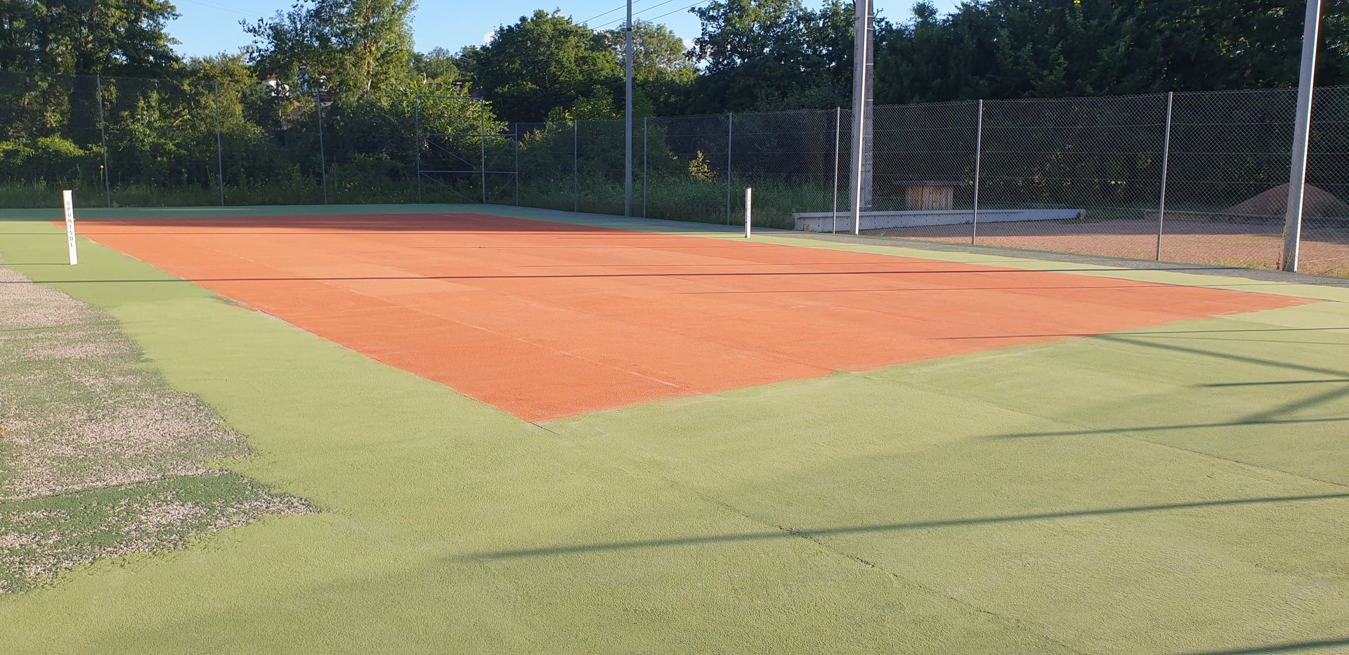 Un court de tennis en terre battue vide, entouré de gazon artificiel vert, avec un filet en arrière-plan.