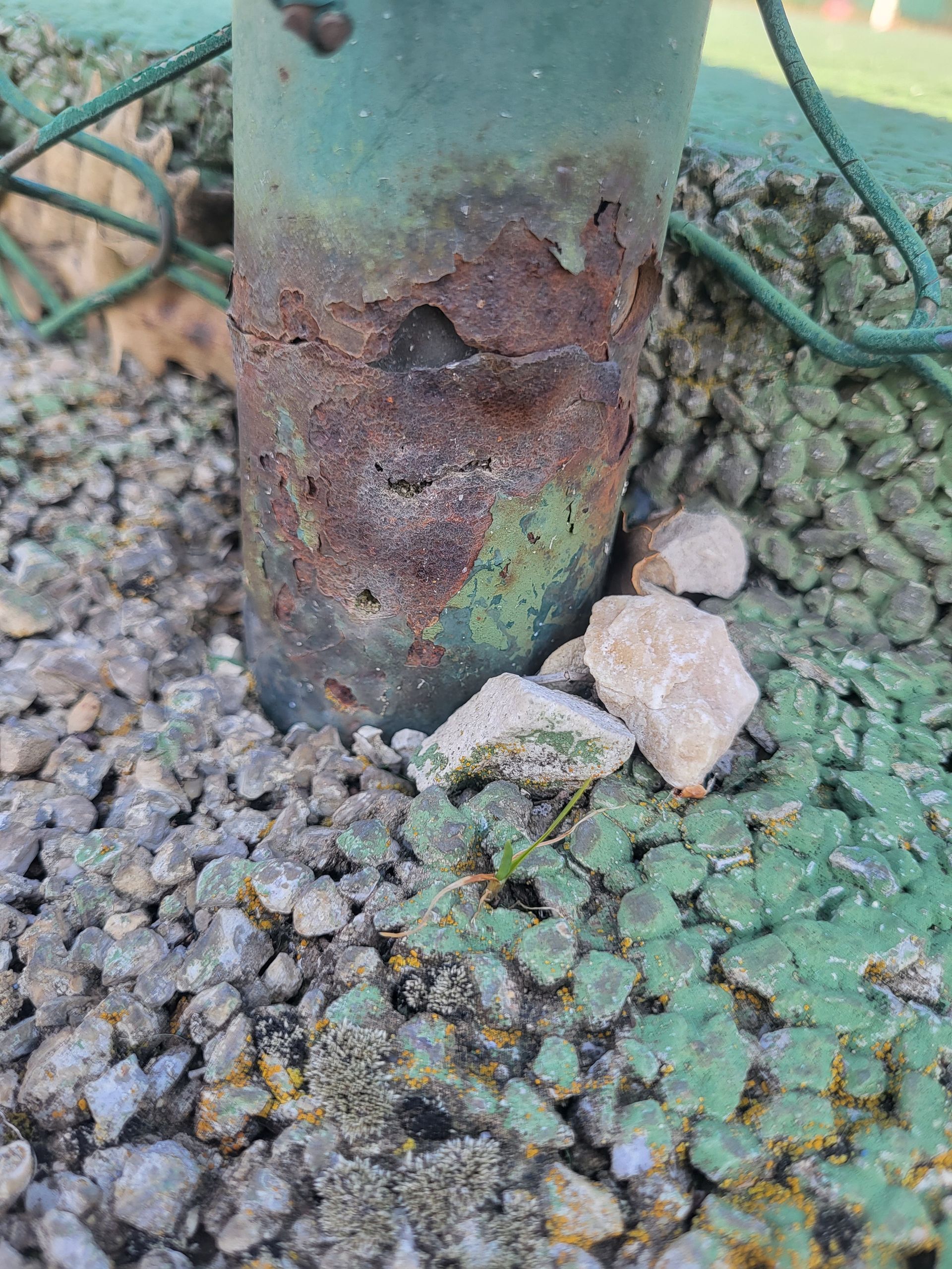 Socle de poteau en métal vert fortement rouillé, entouré de gravier, servant probablement à soutenir une clôture.