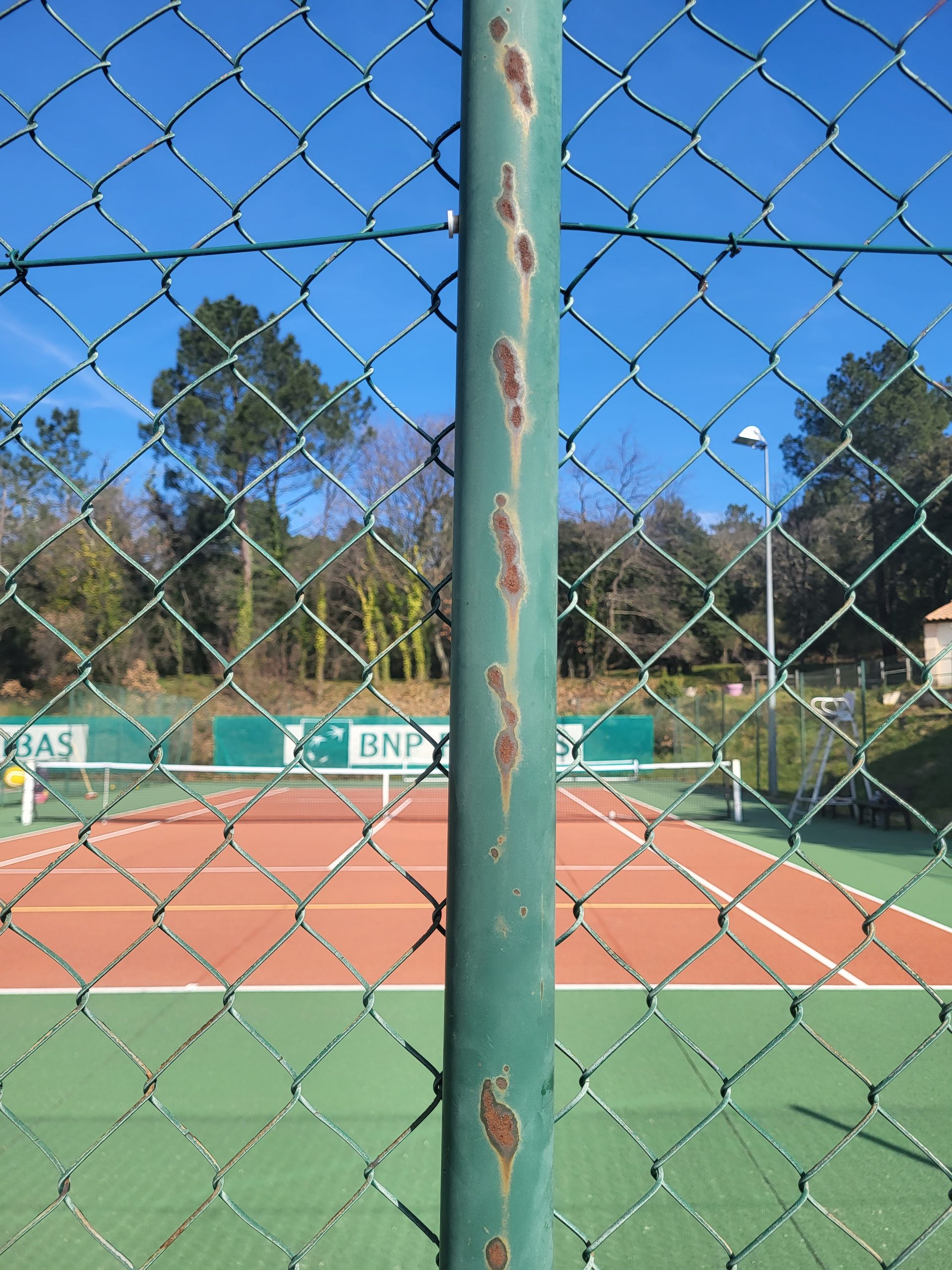 Court de tennis délimité par une clôture en grillage vert, ciel bleu, arbres verts et un poteau métallique usé.