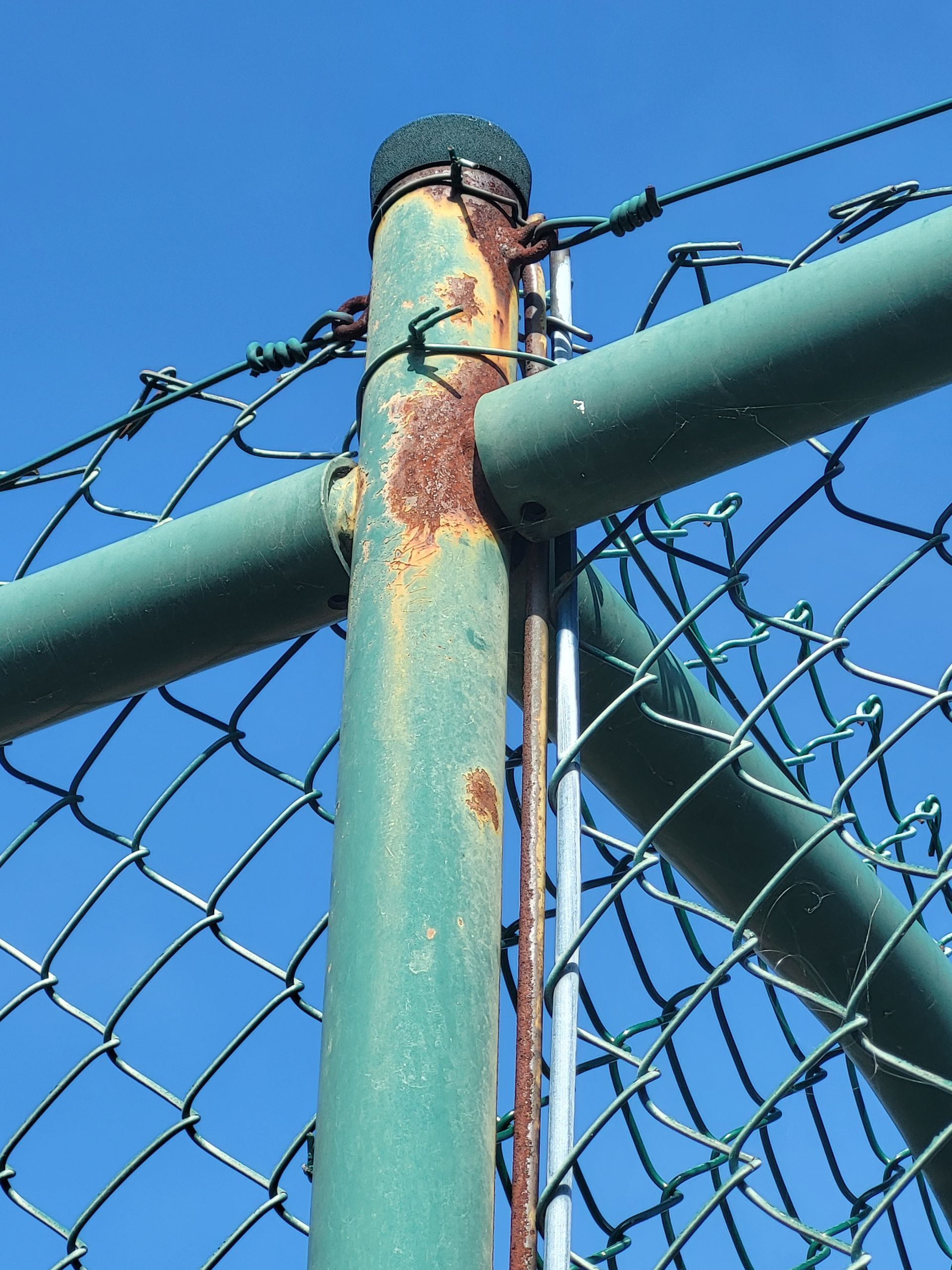 Poteau de clôture en métal vert rouillé et grillage, sur fond de ciel bleu.