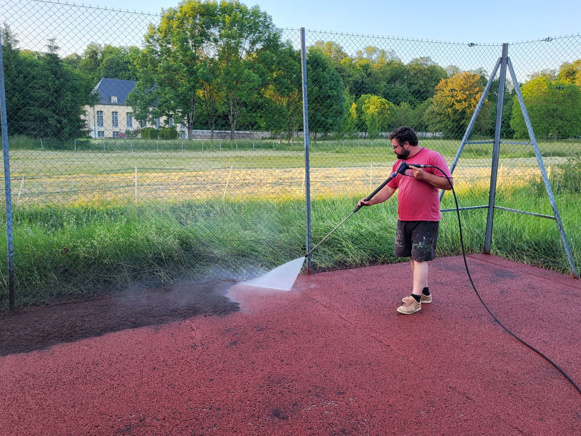 Un homme nettoie une surface rouge au nettoyeur haute pression, près d'un champ herbeux et d'un bâtiment.