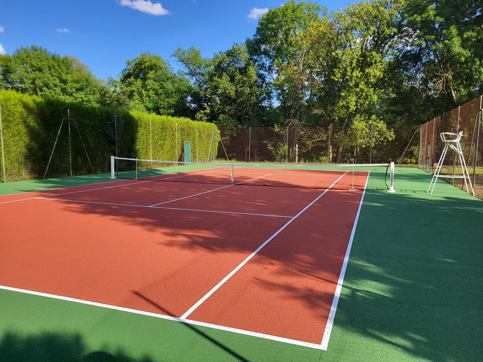 Court de tennis en terre battue rouge avec des lignes blanches, entouré de gazon artificiel vert et d'arbres.