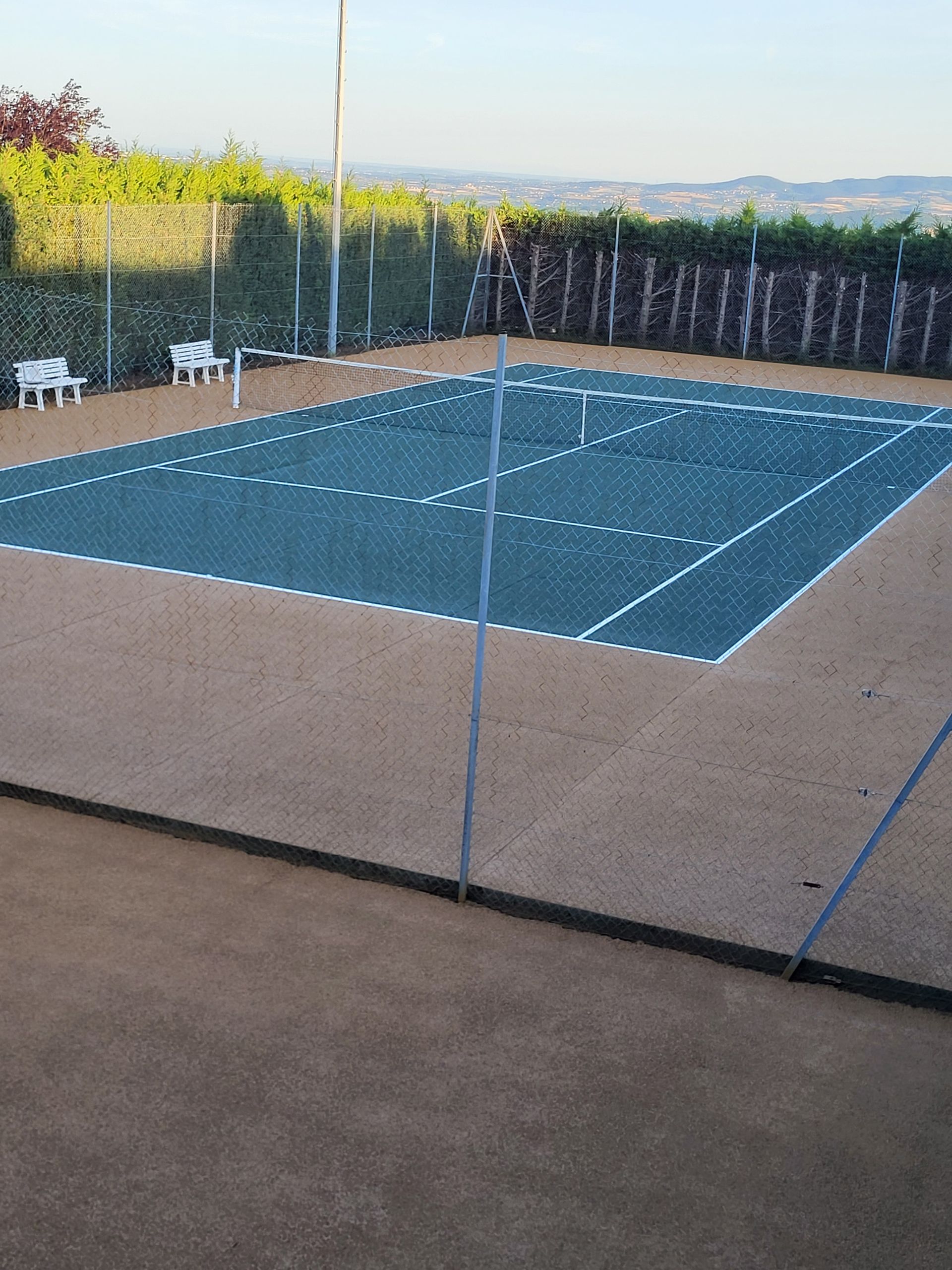 Court de tennis avec surface de jeu bleue et bordures beiges, avec arbres et ciel en arrière-plan.