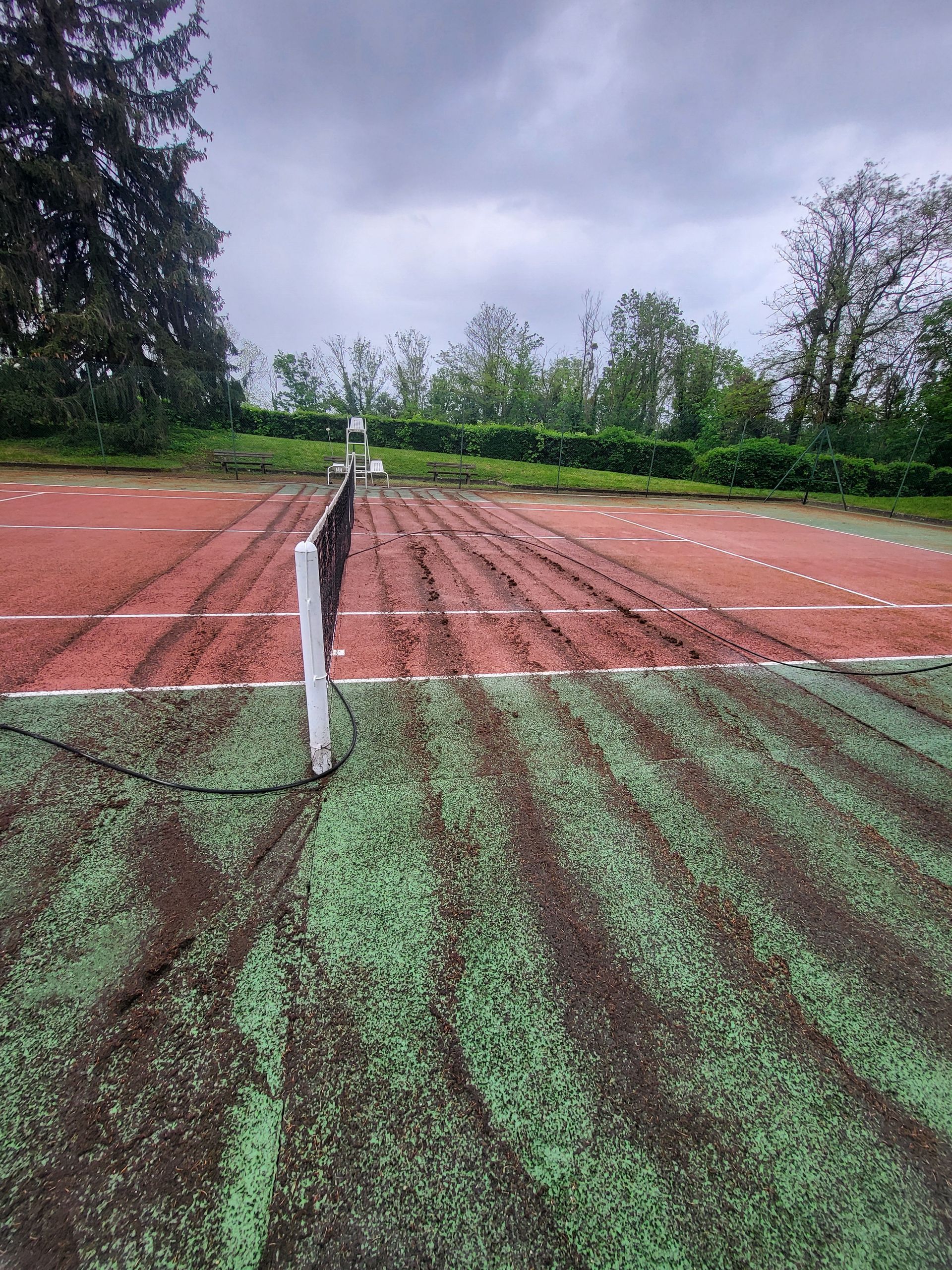 Court de tennis boueux avec des surfaces de jeu vertes et rouges, un filet et un ciel couvert.