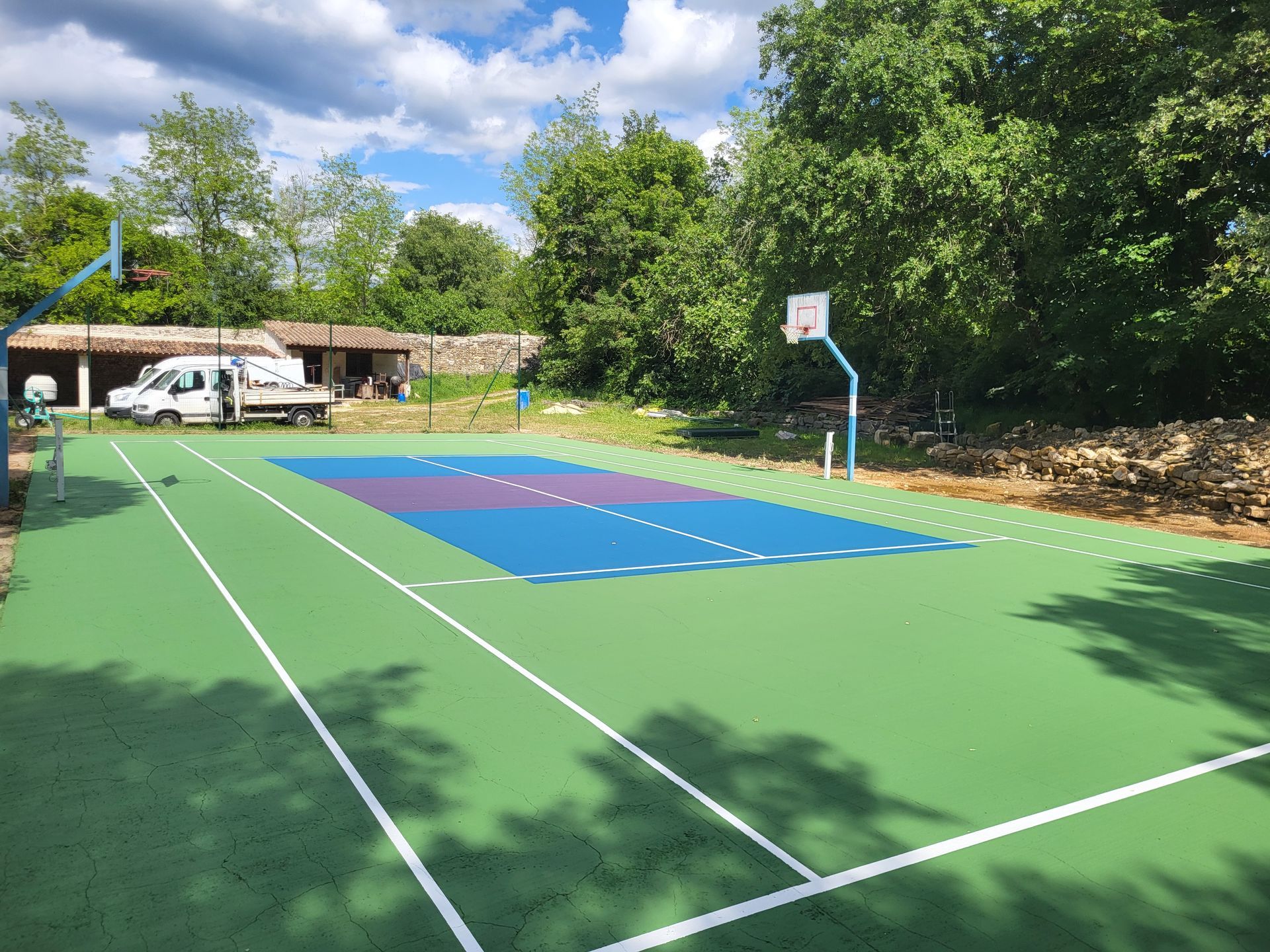Terrain de basket-ball extérieur avec des surfaces de jeu vertes, bleues et violettes, avec deux paniers, devant des arbres.