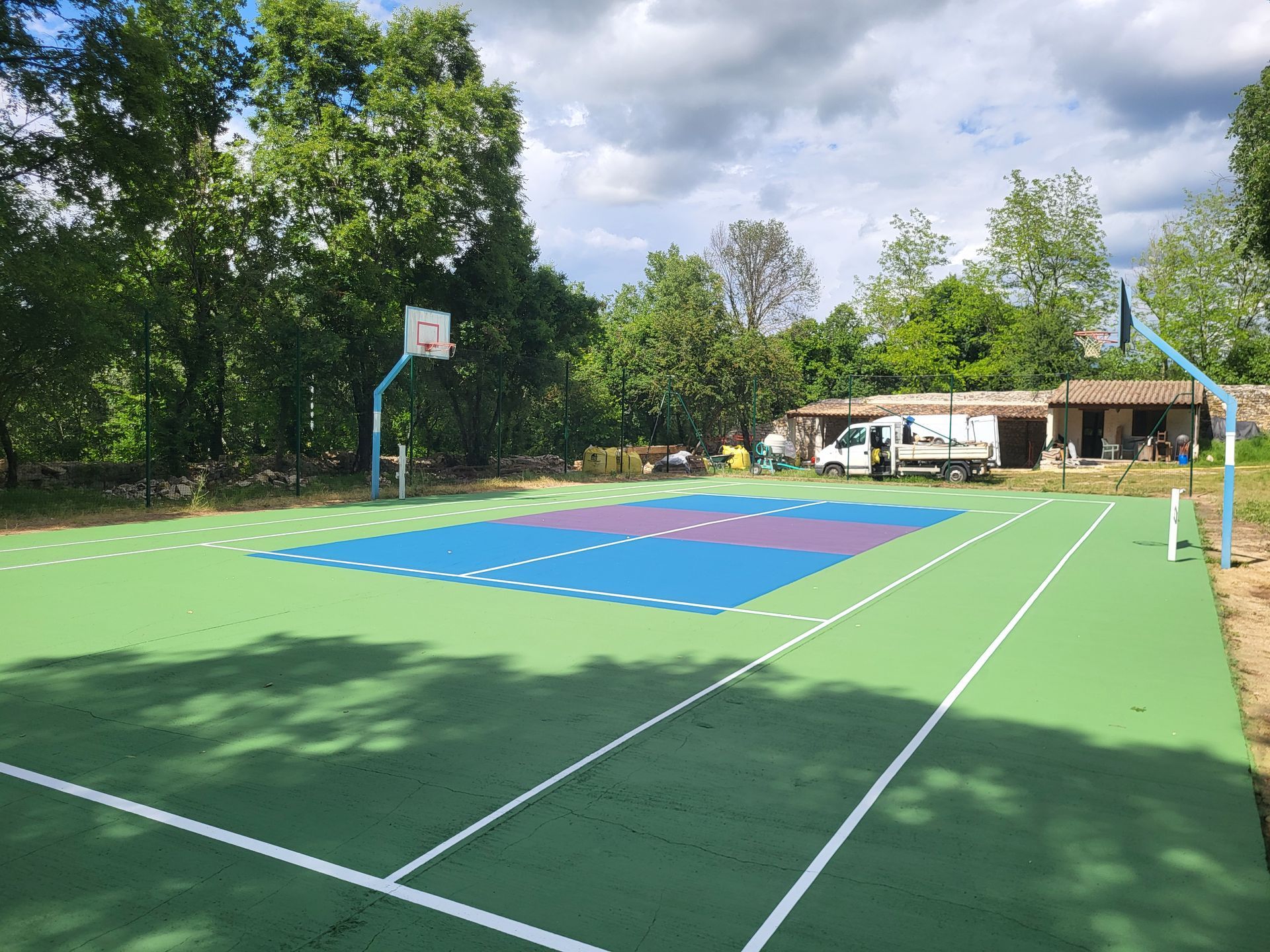 Terrain de basket-ball, vert et bleu, avec panneau, arbres et petit bâtiment en arrière-plan.