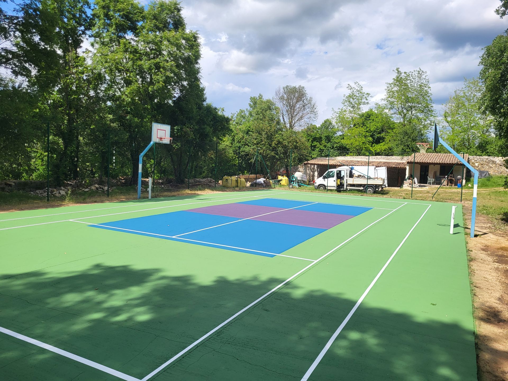 Terrain de basket-ball extérieur avec surfaces vertes et bleues, paniers et arbres en arrière-plan.