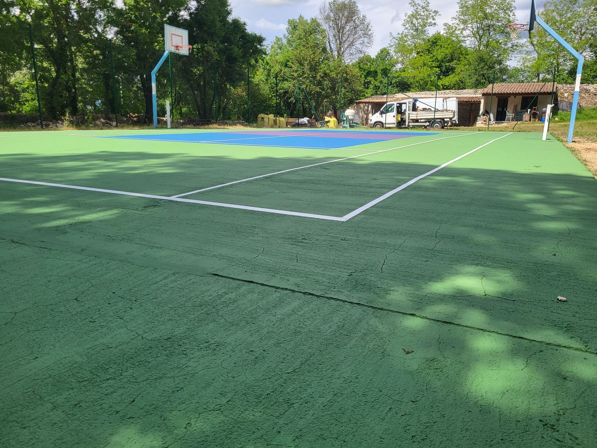 Terrain de basket-ball vert et bleu avec des lignes blanches, entouré d'arbres et d'un petit bâtiment.