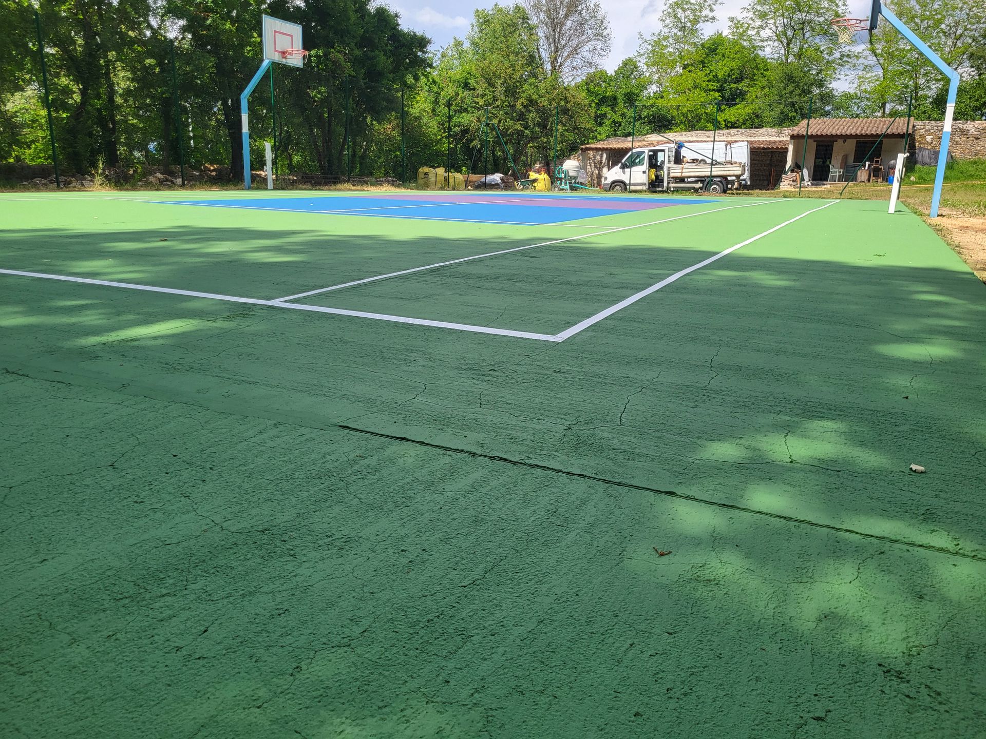 Terrain de basket-ball extérieur avec surfaces vertes et bleues, arbres et lignes blanches.