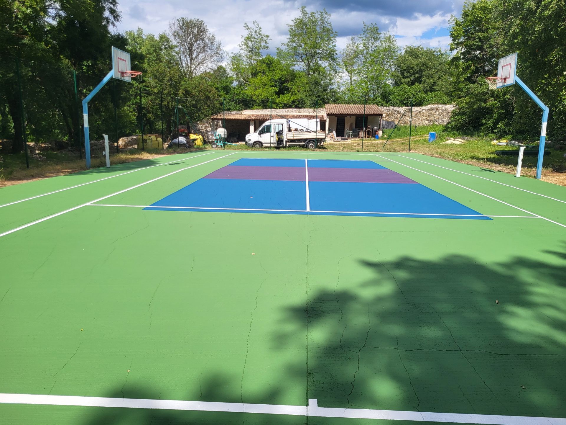Terrain de basket avec une surface de jeu verte et bleue, des paniers et un petit bâtiment en arrière-plan.