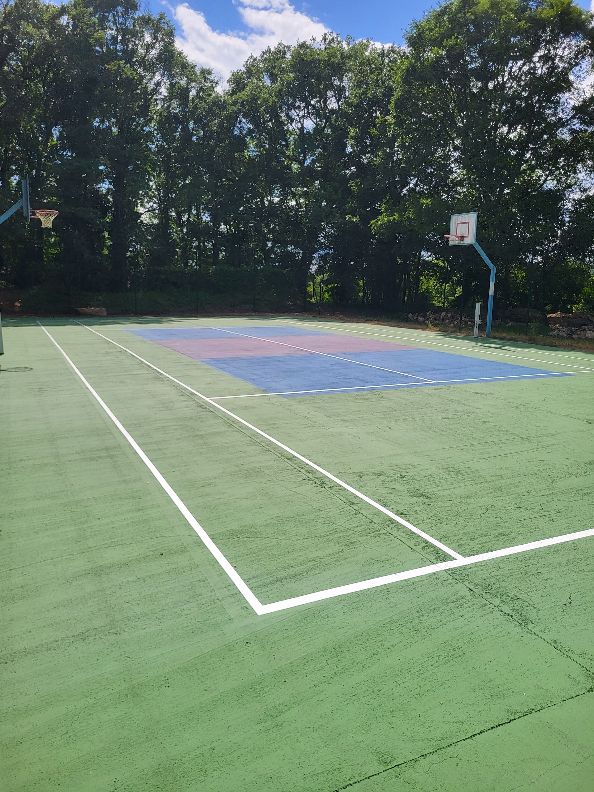 Terrain de basket avec des surfaces bleues et vertes, deux paniers et des arbres environnants sous un ciel bleu.