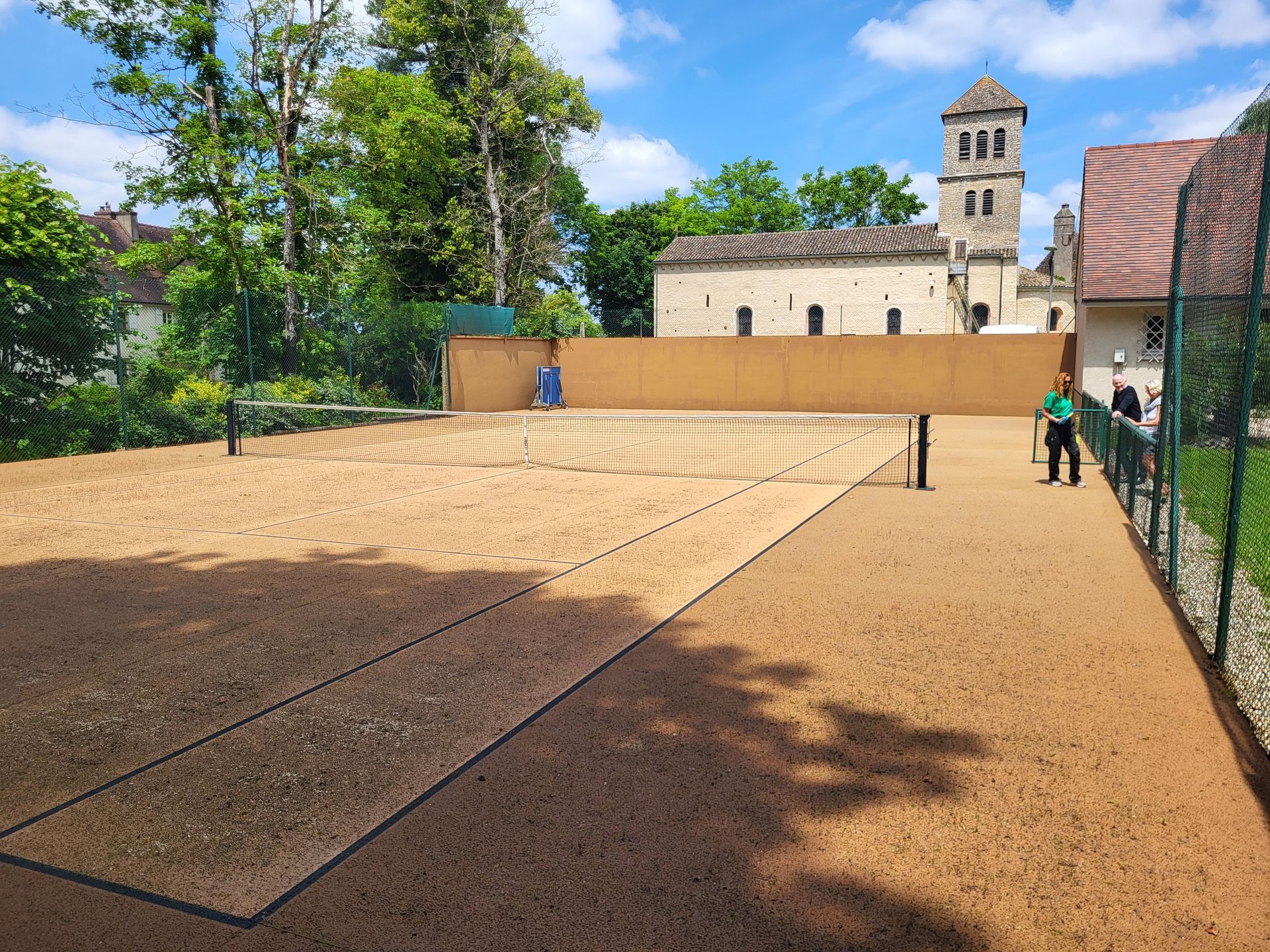Court de tennis avec une personne, surface beige, filet, église en arrière-plan.
