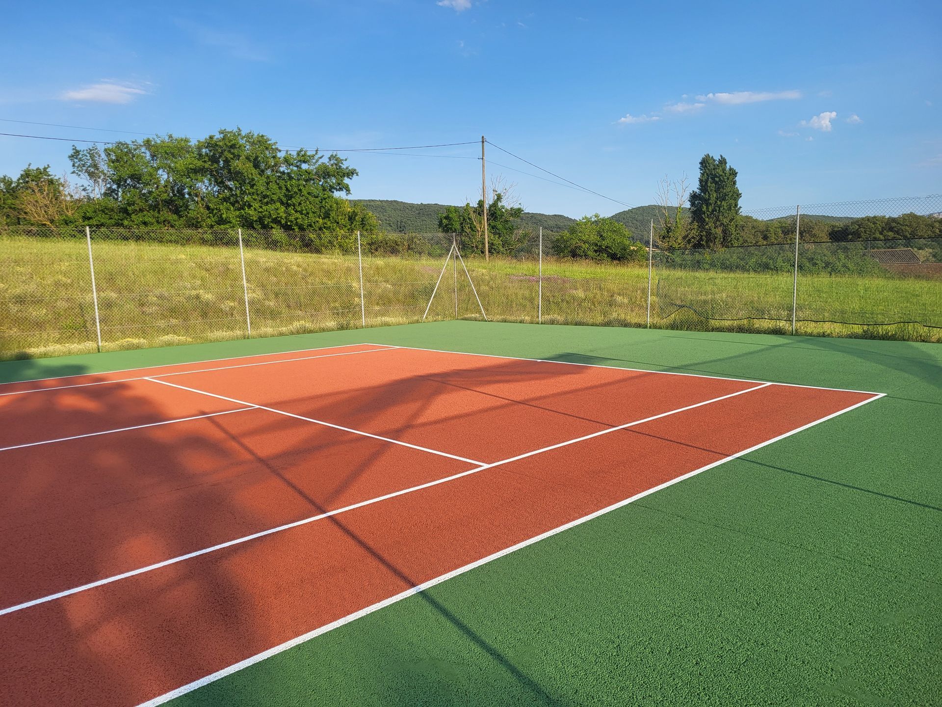 Terrain extérieur rouge et vert avec lignes blanches, clôture, arbres et ciel bleu.