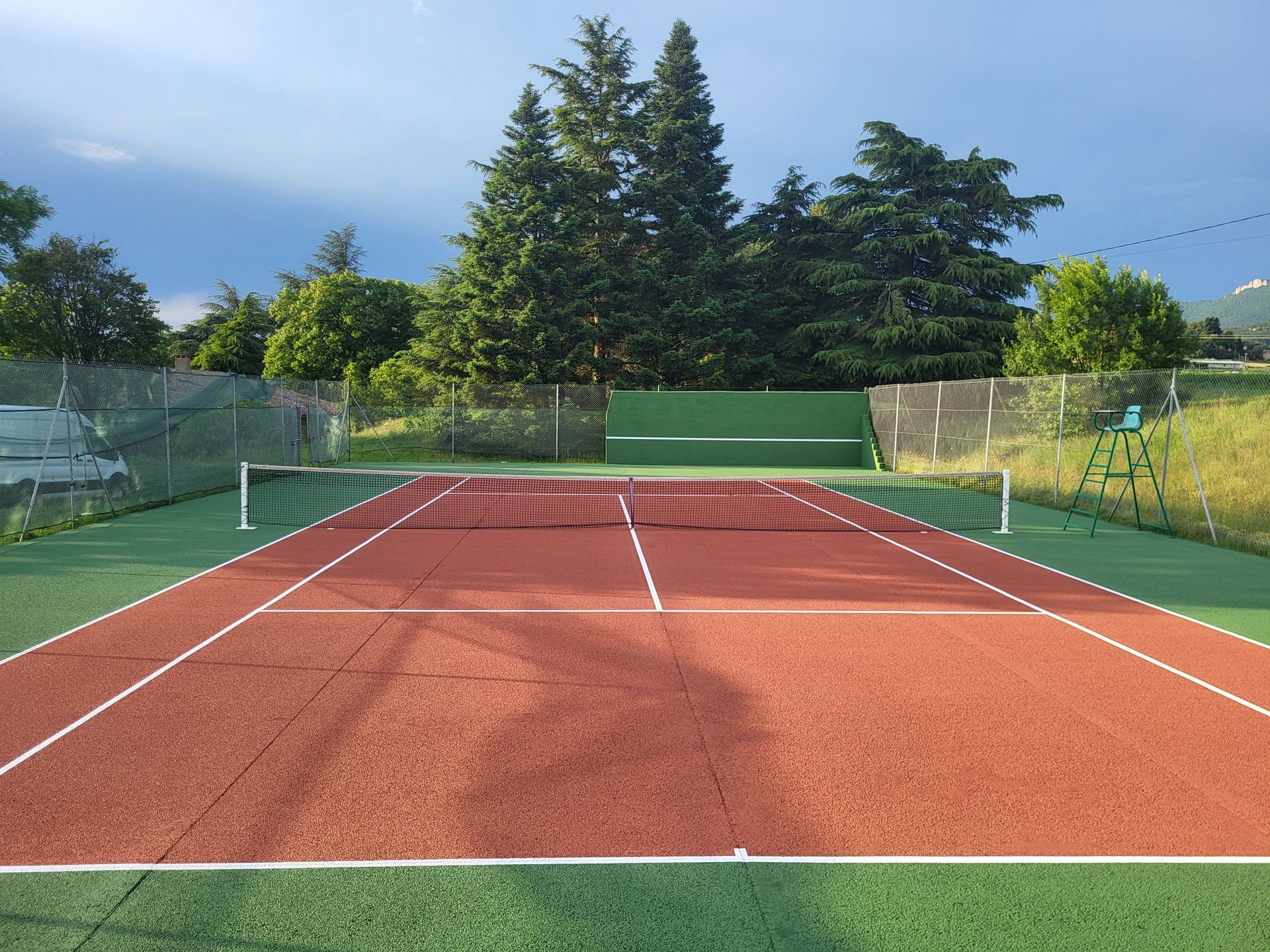 Court de tennis avec surface en terre battue rouge, lignes blanches, zone environnante verte et filet.