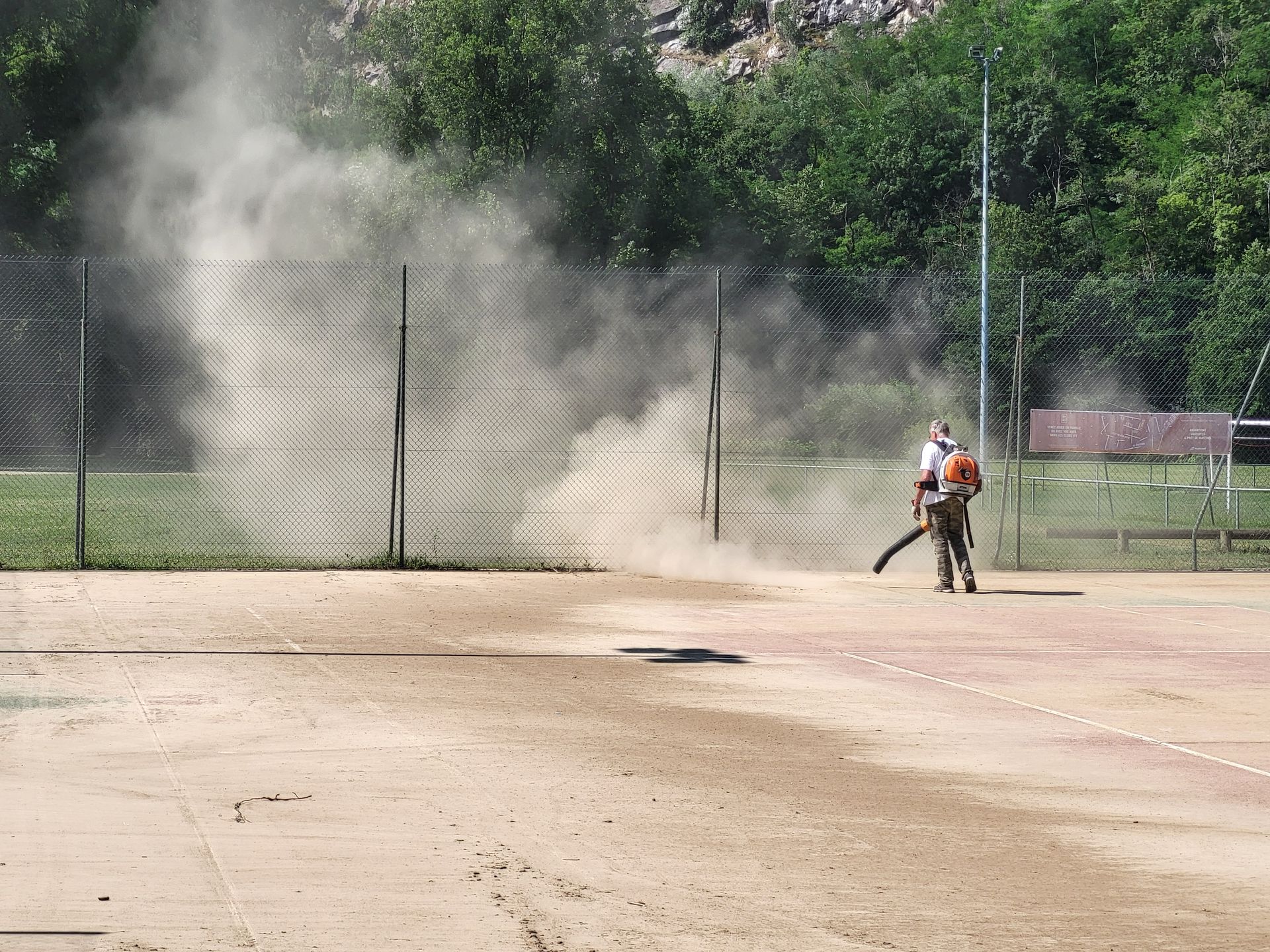 Un homme souffle la poussière sur un court de tennis en terre battue, près d'une clôture en grillage et d'arbres.