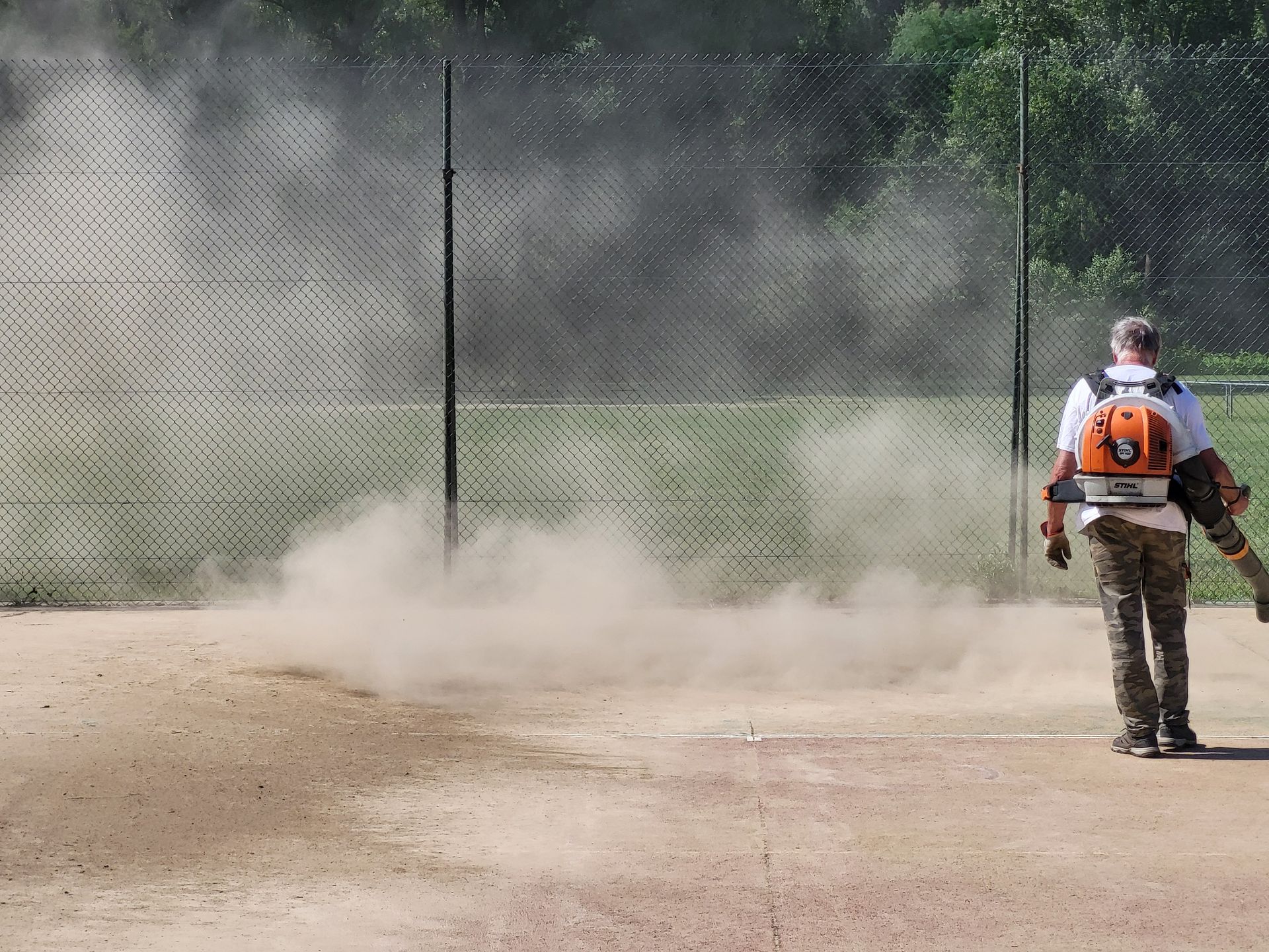 Une personne utilise un souffleur de feuilles, soulevant de la poussière sur un terrain de baseball en terre battue près d'une clôture.