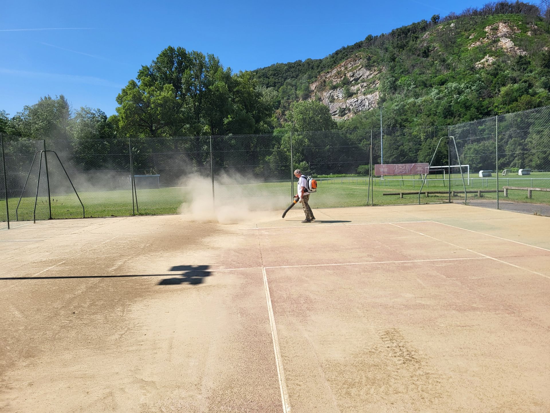 Une personne manipule une machine sur un terrain sablonneux, soulevant des nuages ​​de poussière. Une clôture et des arbres se dressent en arrière-plan, avec des montagnes à l'horizon.