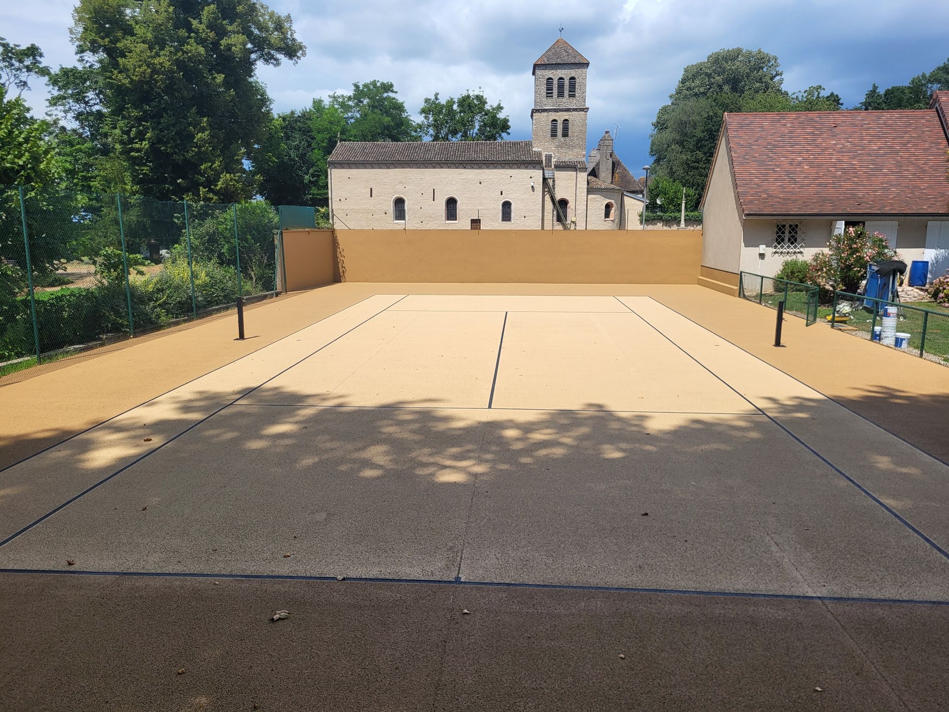 Une cour de gravier délimitée par des lignes, bordée de bâtiments, d'arbres et d'une église au loin.