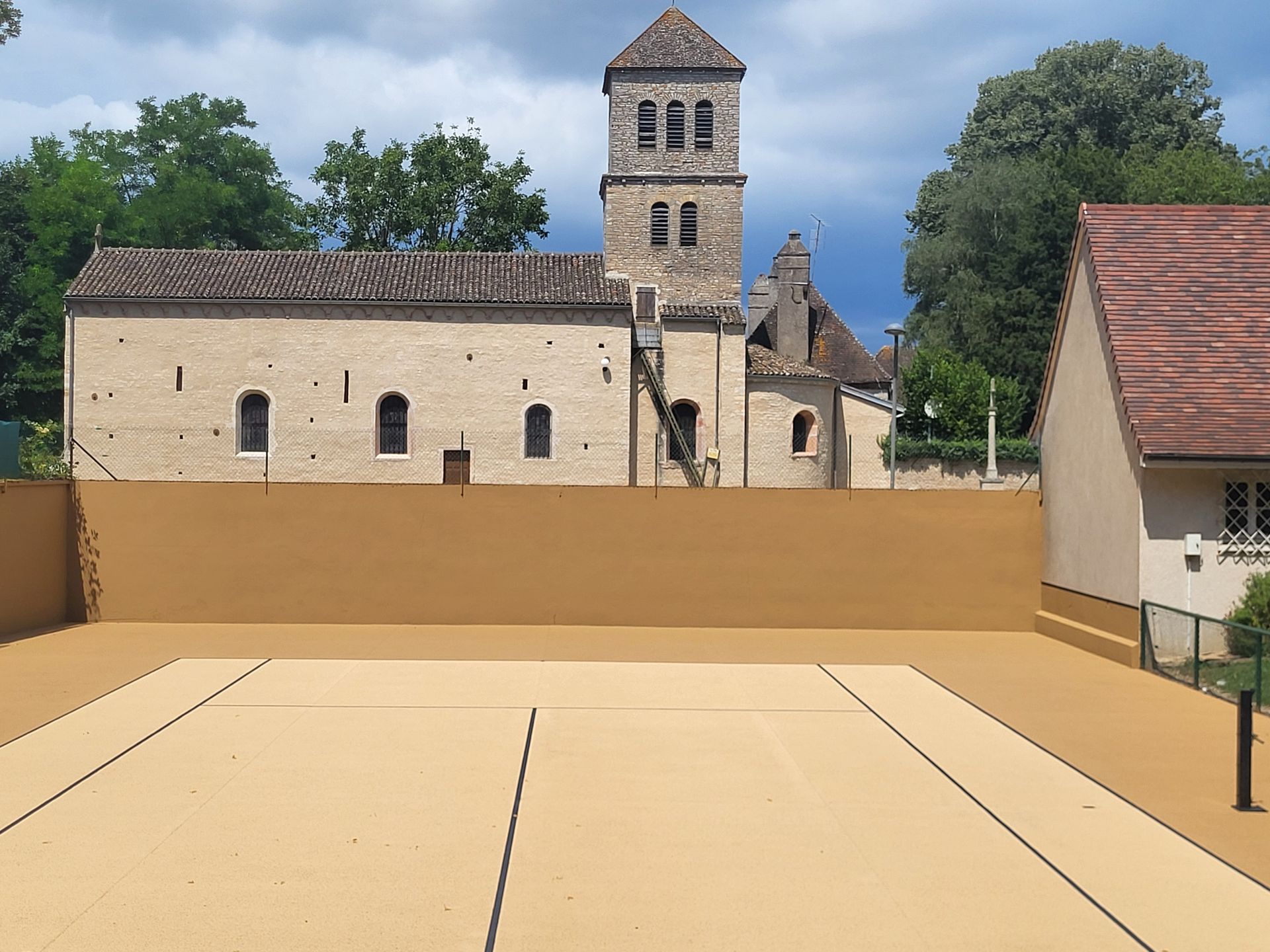 Une cour de fronton beige avec une église en arrière-plan. Un muret beige et un petit bâtiment se trouvent de part et d'autre.