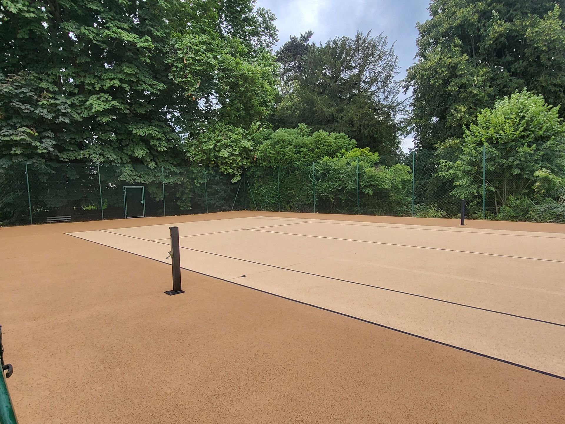 Court de tennis en terre battue avec filet et arbres en arrière-plan sous un ciel nuageux.