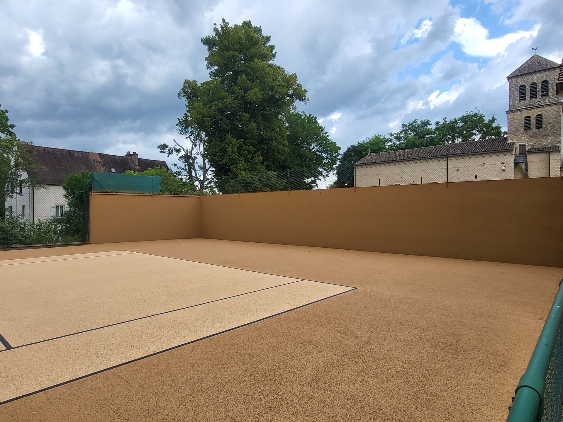 Terrain de pétanque extérieur avec surface beige, murs marron et église en arrière-plan.