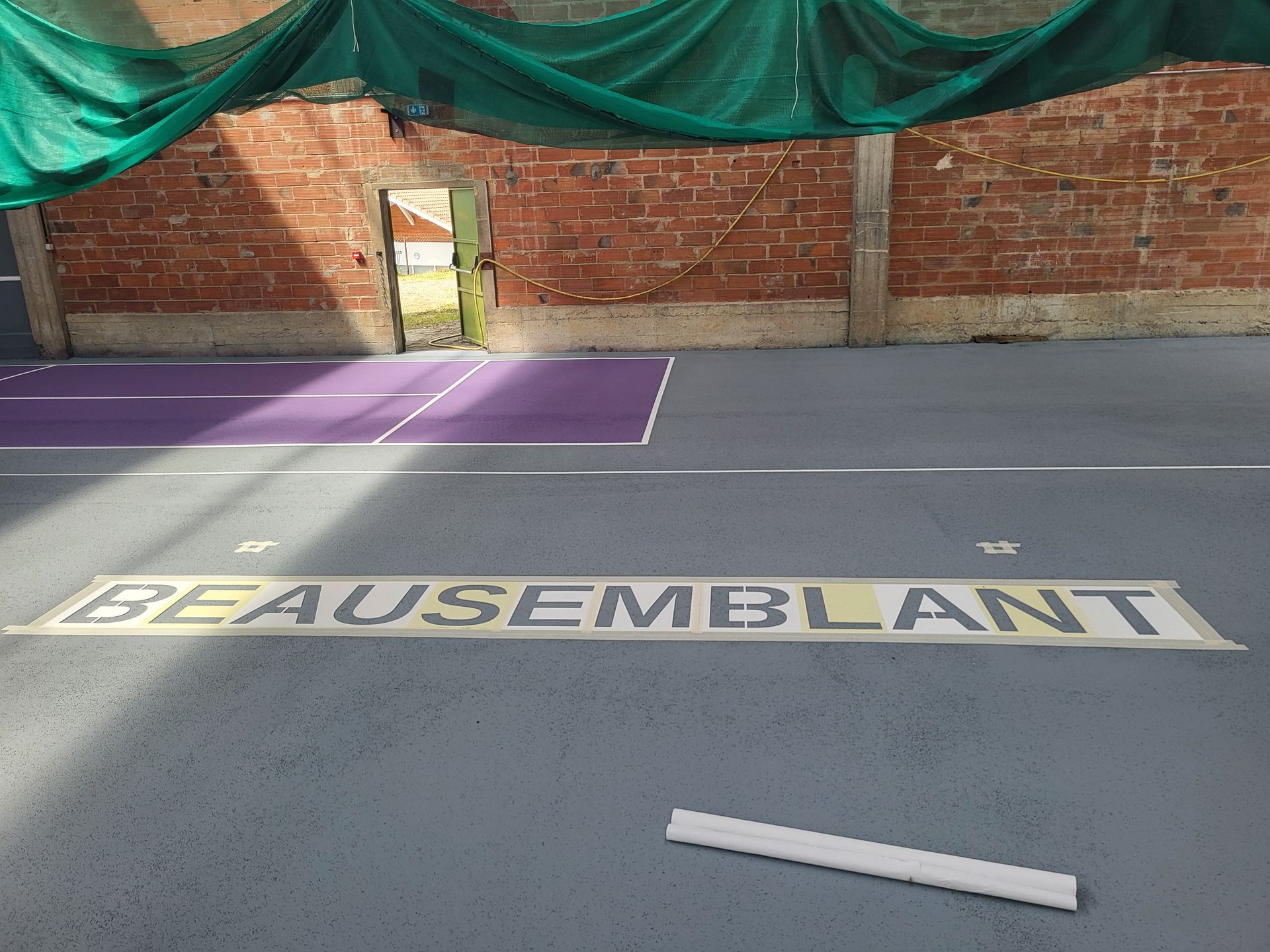 Terrain de sport intérieur avec l'inscription BEAUSEMBLANT au pochoir sur le sol ; mur de briques rouges en arrière-plan.