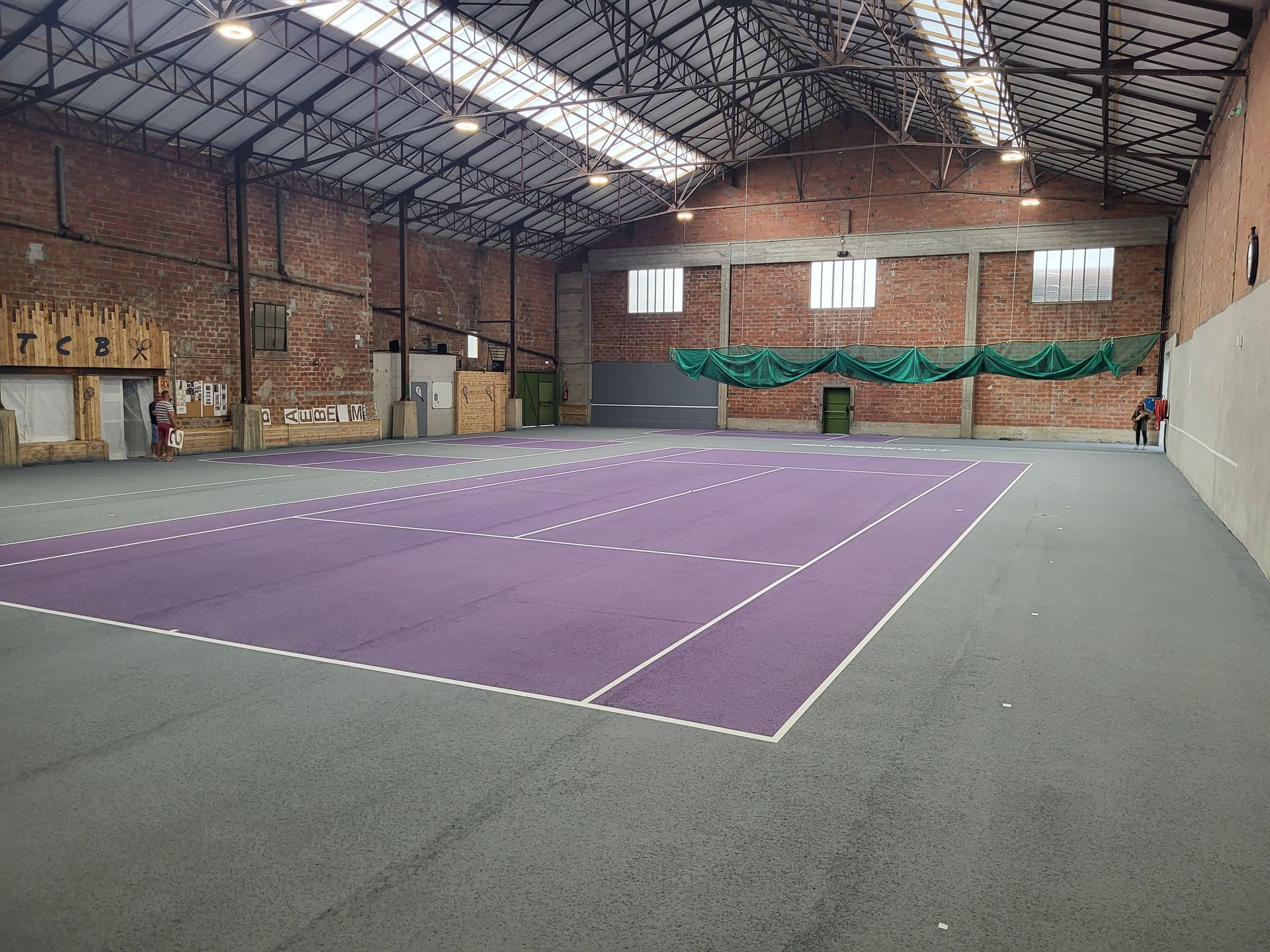 Terrain de sport intérieur avec surface violette, lignes marquées, murs en briques et haut plafond.