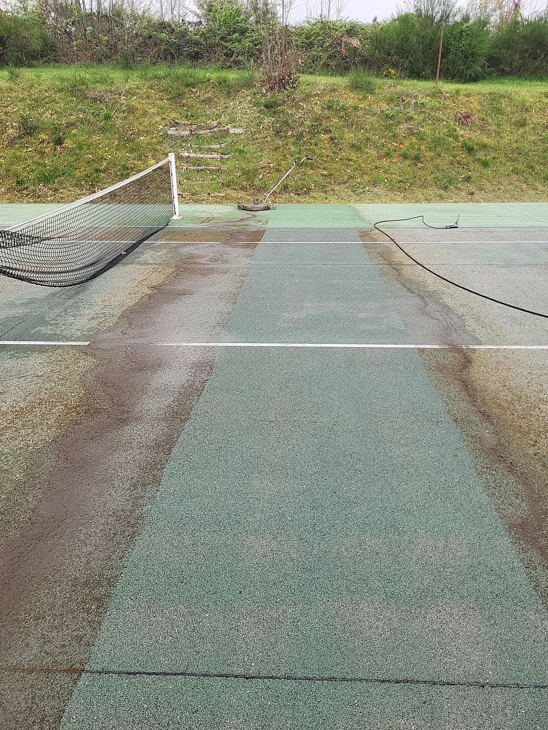 Court de tennis vert et marron avec filet, vu du ciel, entouré d'herbe et de feuillage.