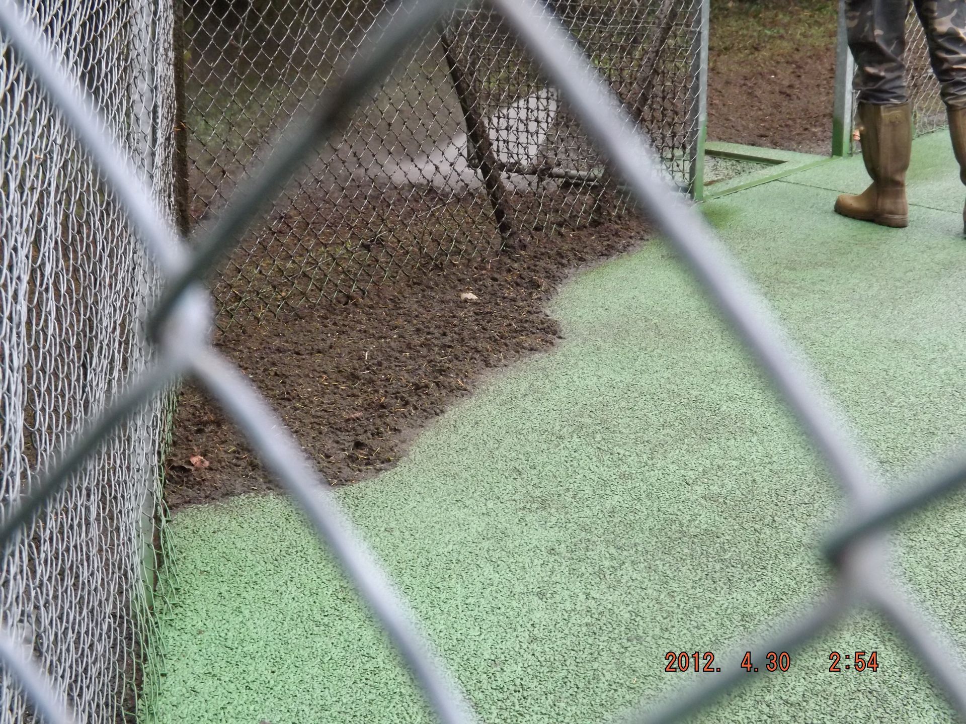 Vue à travers un grillage d'une zone inondée avec un sol vert et une personne en bottes se tenant à proximité.