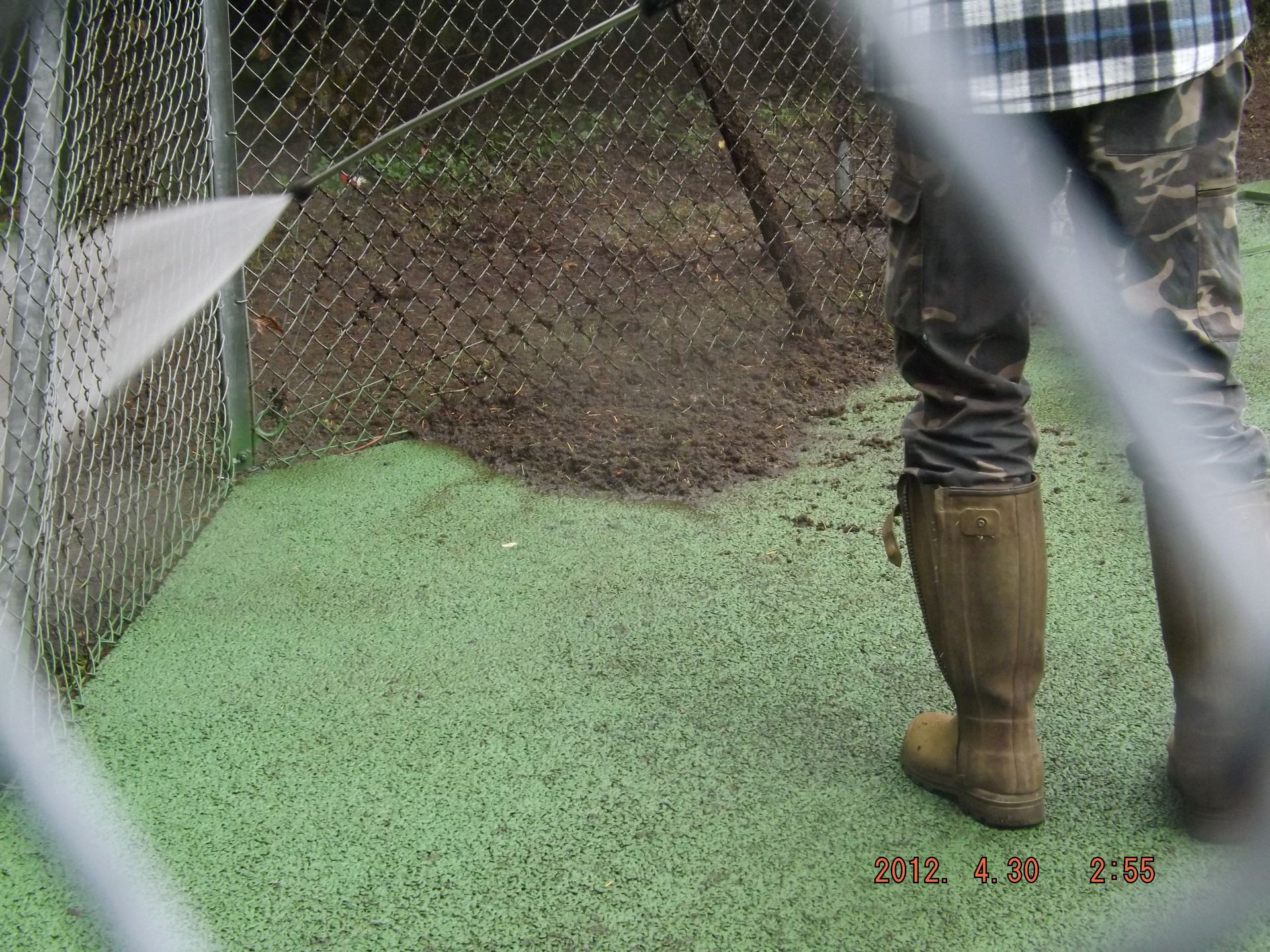 Une personne arrosant une clôture en grillage et une surface verte, portant des bottes en caoutchouc et un pantalon de camouflage.