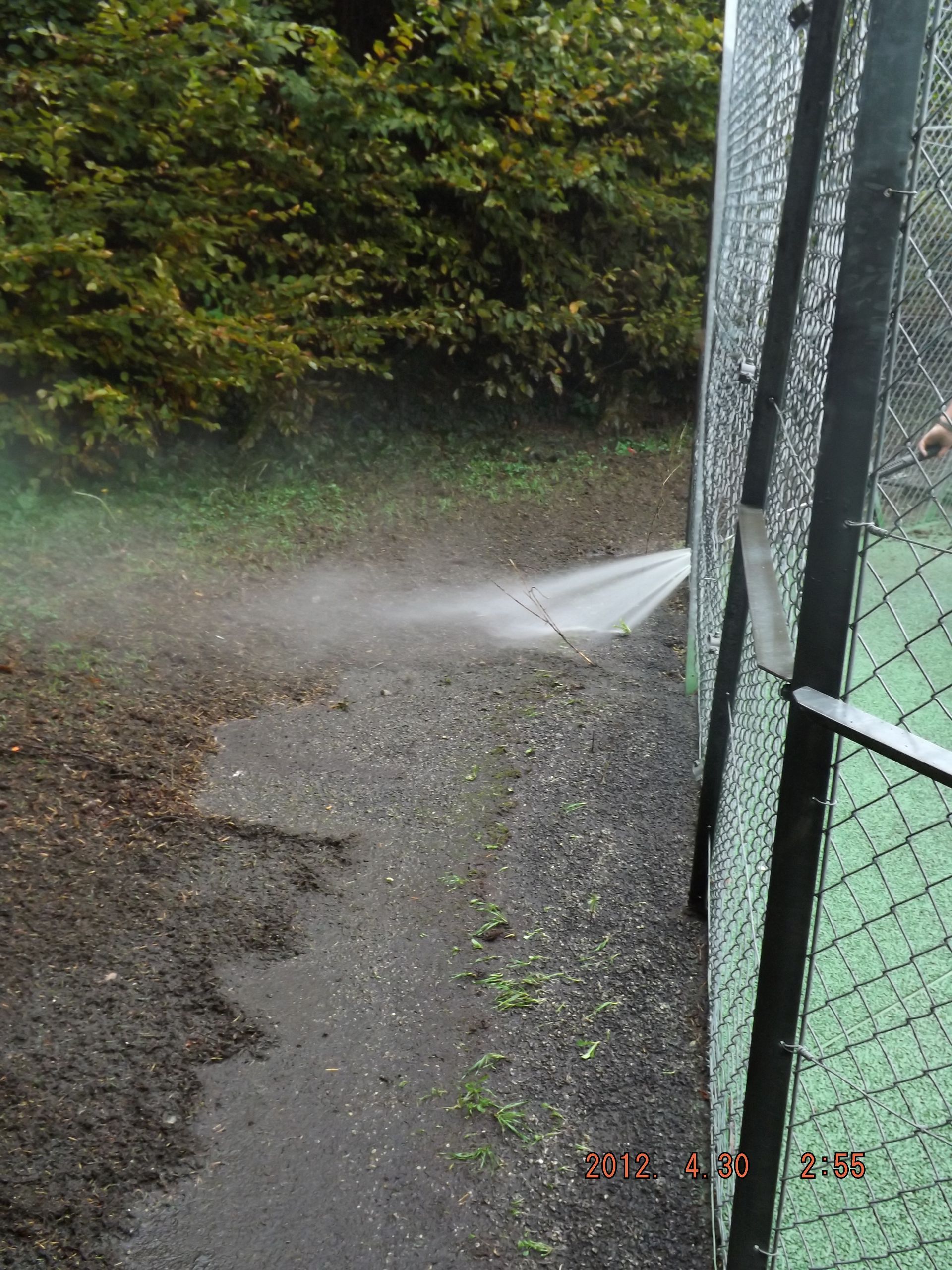 De l'eau jaillit d'un tuyau cassé près de la clôture d'un court de tennis et d'un chemin de gravier.