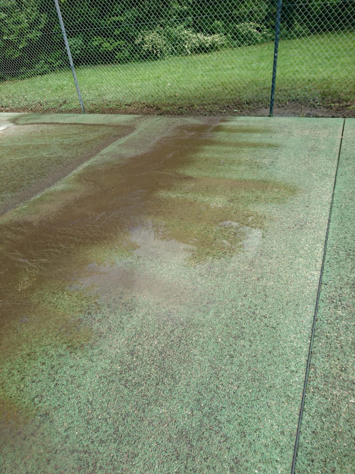 Zone humide et tachée de brun sur un court de tennis vert, près d'une clôture en grillage et de l'herbe.