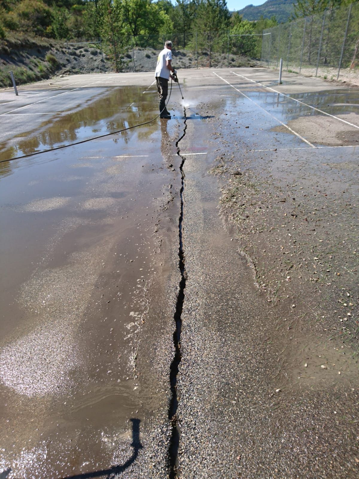 Un homme nettoie à haute pression une surface asphaltée fissurée et recouverte d'eau, en extérieur.